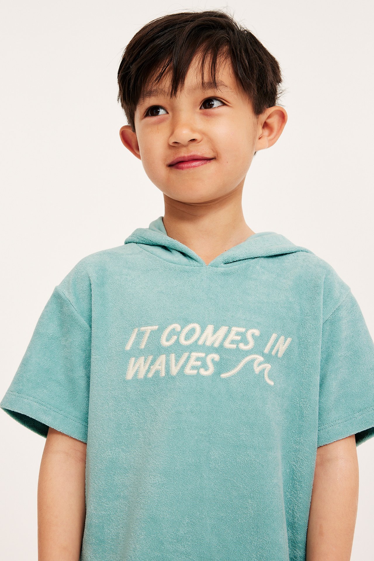 Frontvy: Barn i oversized ljusgrön kortärmad frottéhoodie med broderad text 'IT COMES IN WAVES'.