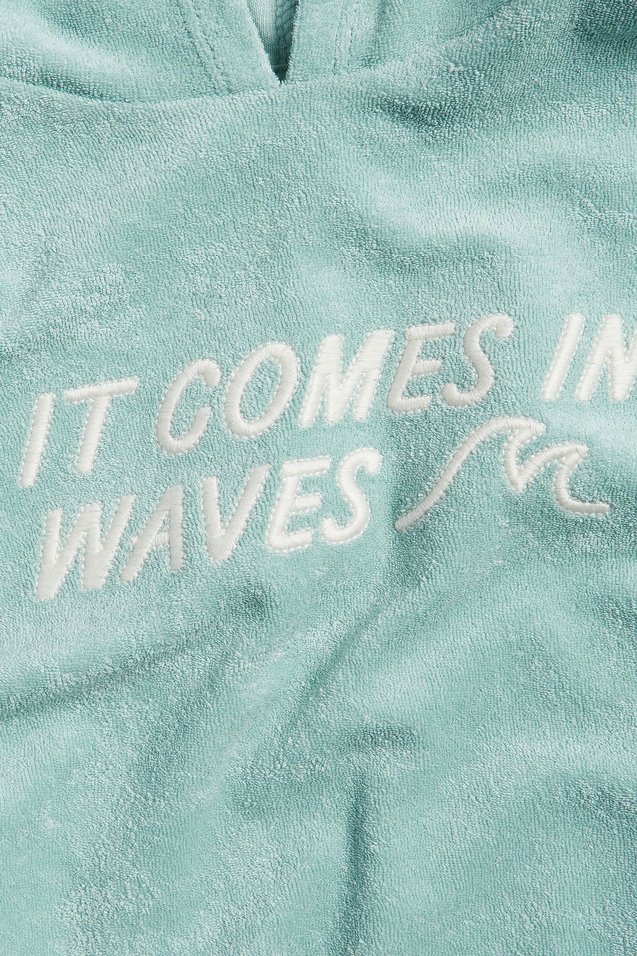 Zbliżenie na jasnozieloną, krótką bluzę z kapturem z frotte z białym haftem "IT COMES IN WAVES".