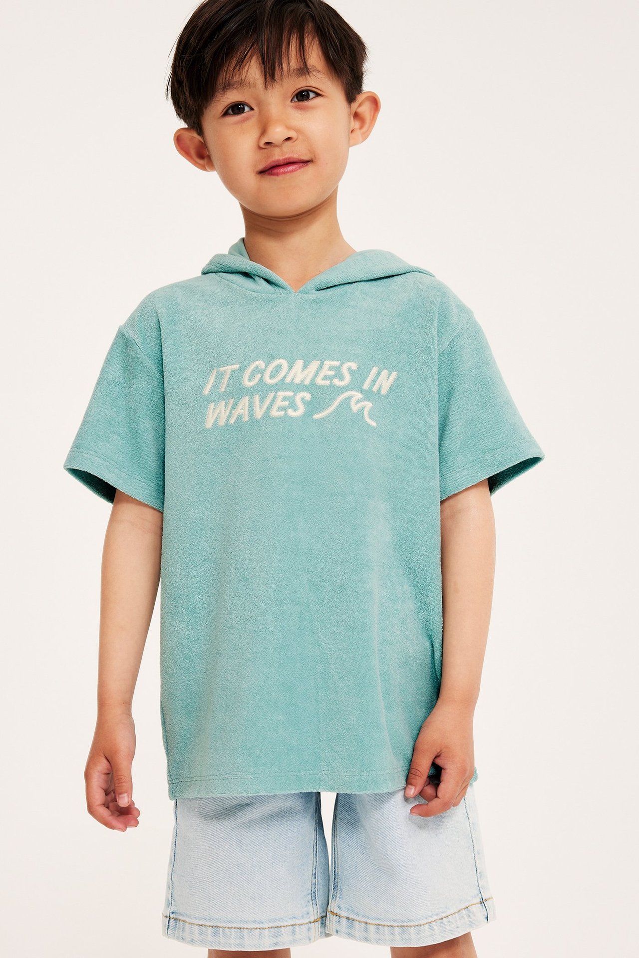 Frontvy av ett barn i en oversized ljusgrön kortärmad frottéhoodie med brodyr och ljusblå denimshorts.