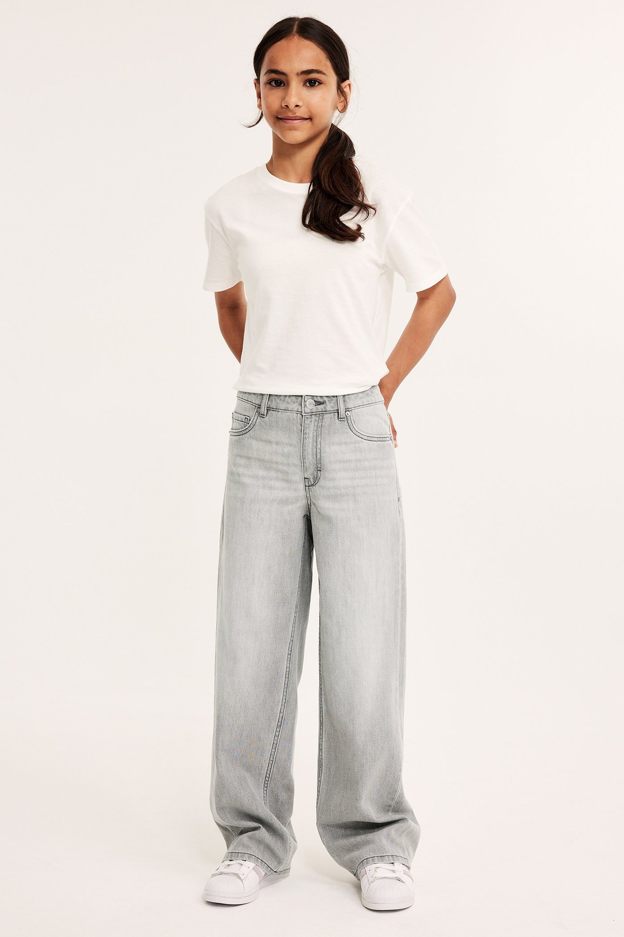 Framifrån: ett barn i gråa, lösa jeans med låg midja, en vit t-shirt och vita sneakers.
