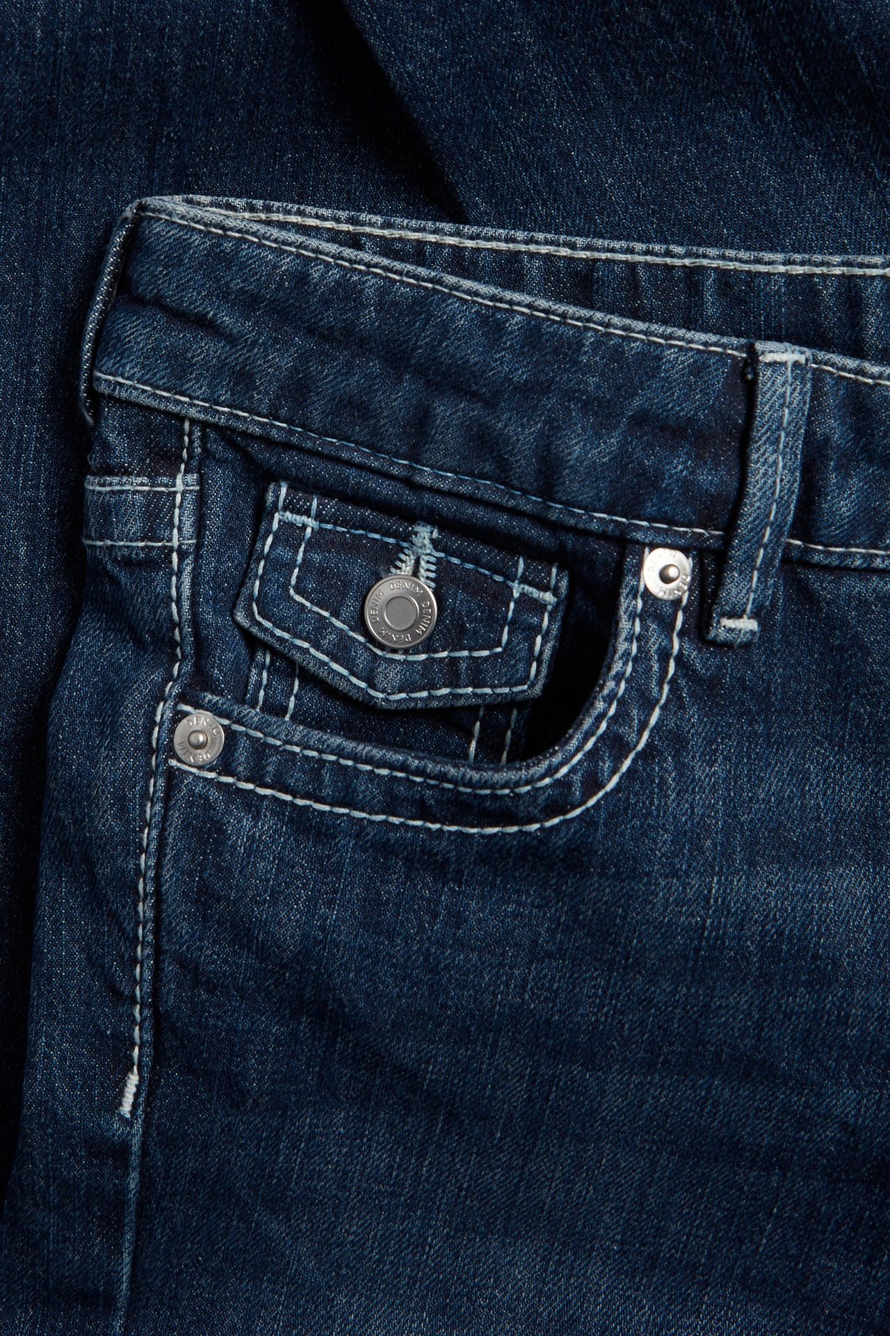Zbliżenie na ciemne jeansy bootcut z jasnymi przeszyciami, przednią kieszenią z klapką i srebrnymi guzikami.