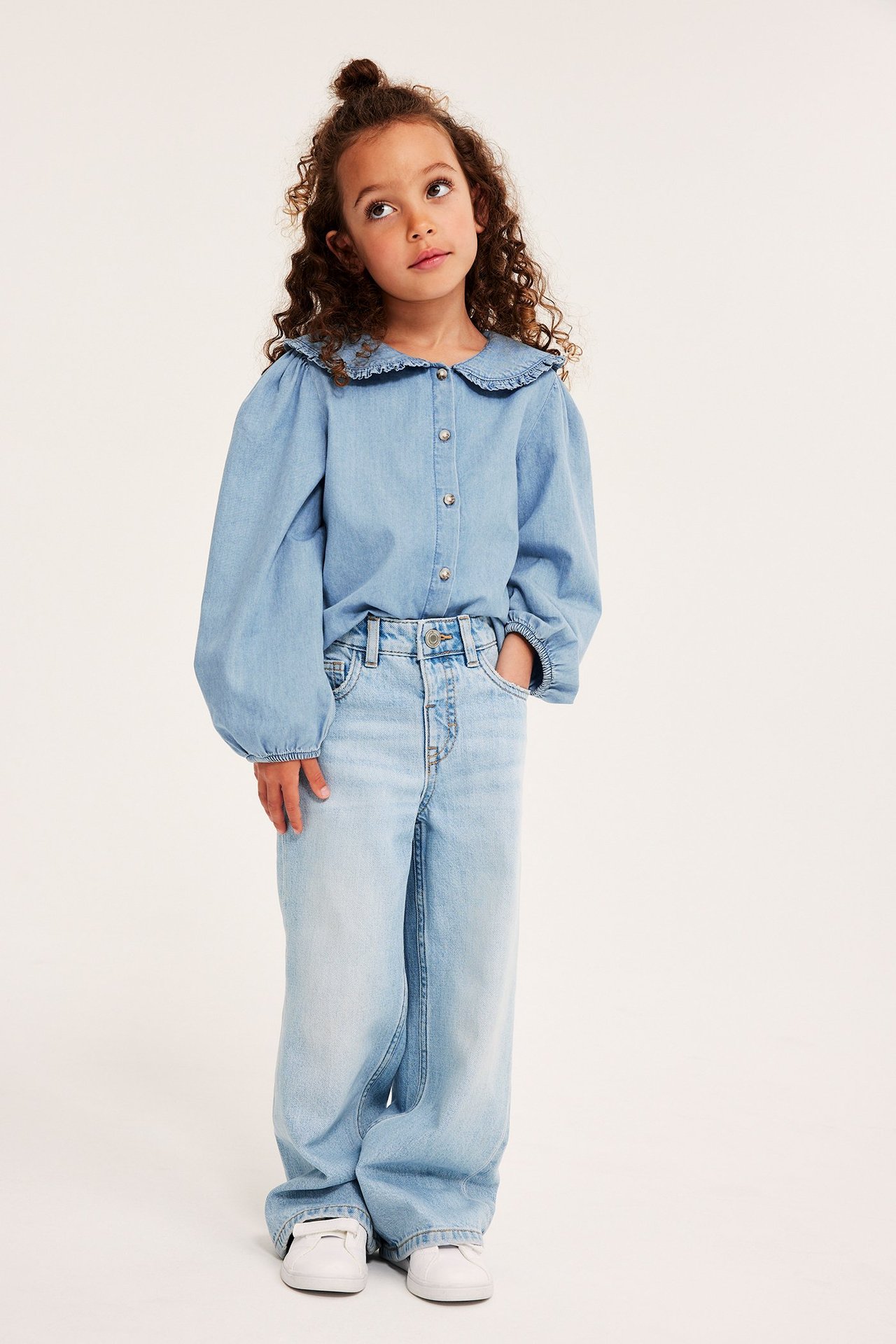 Framifrån: Barn i ljusblå denimblus med volangkrage, ljusa vida jeans och vita sneakers.