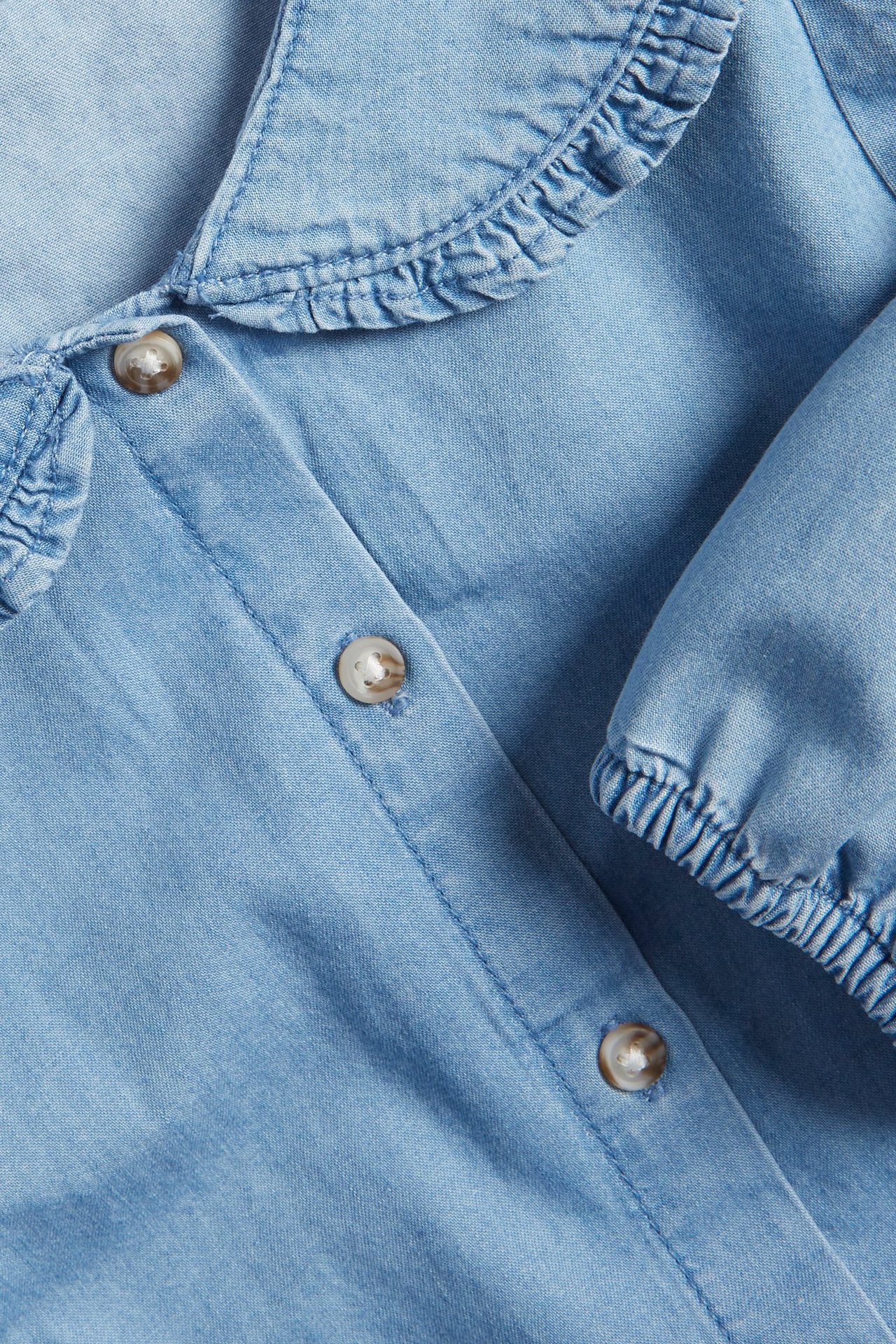 Zbliżenie jasnoniebieskiej bluzki dżinsowej z falbankowym kołnierzem, guzikami z przodu i elastycznym mankietem.