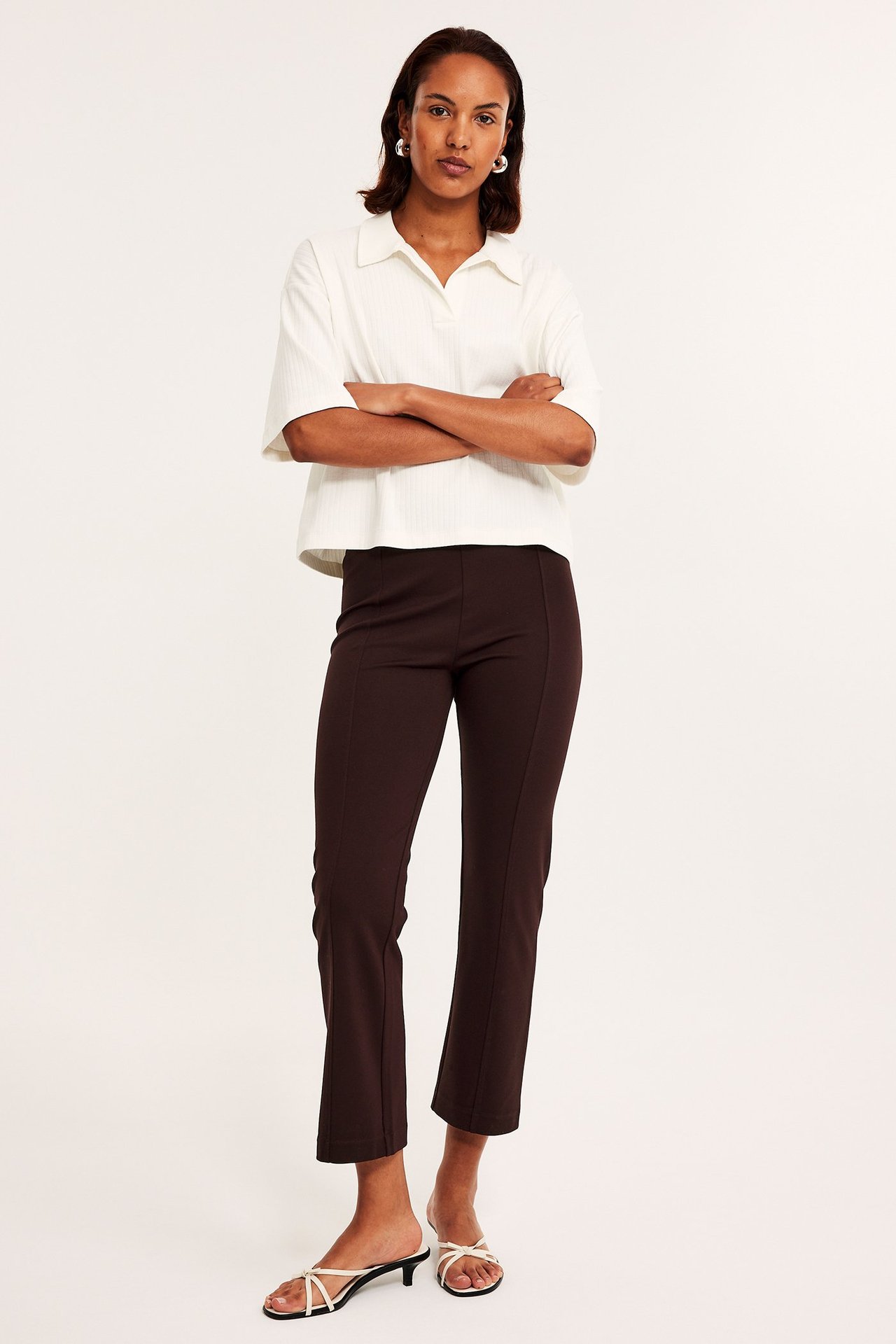 Kobieta z przodu: winnoczerwone spodnie dżersejowe kickflare, kremowa koszulka polo i białe sandały.