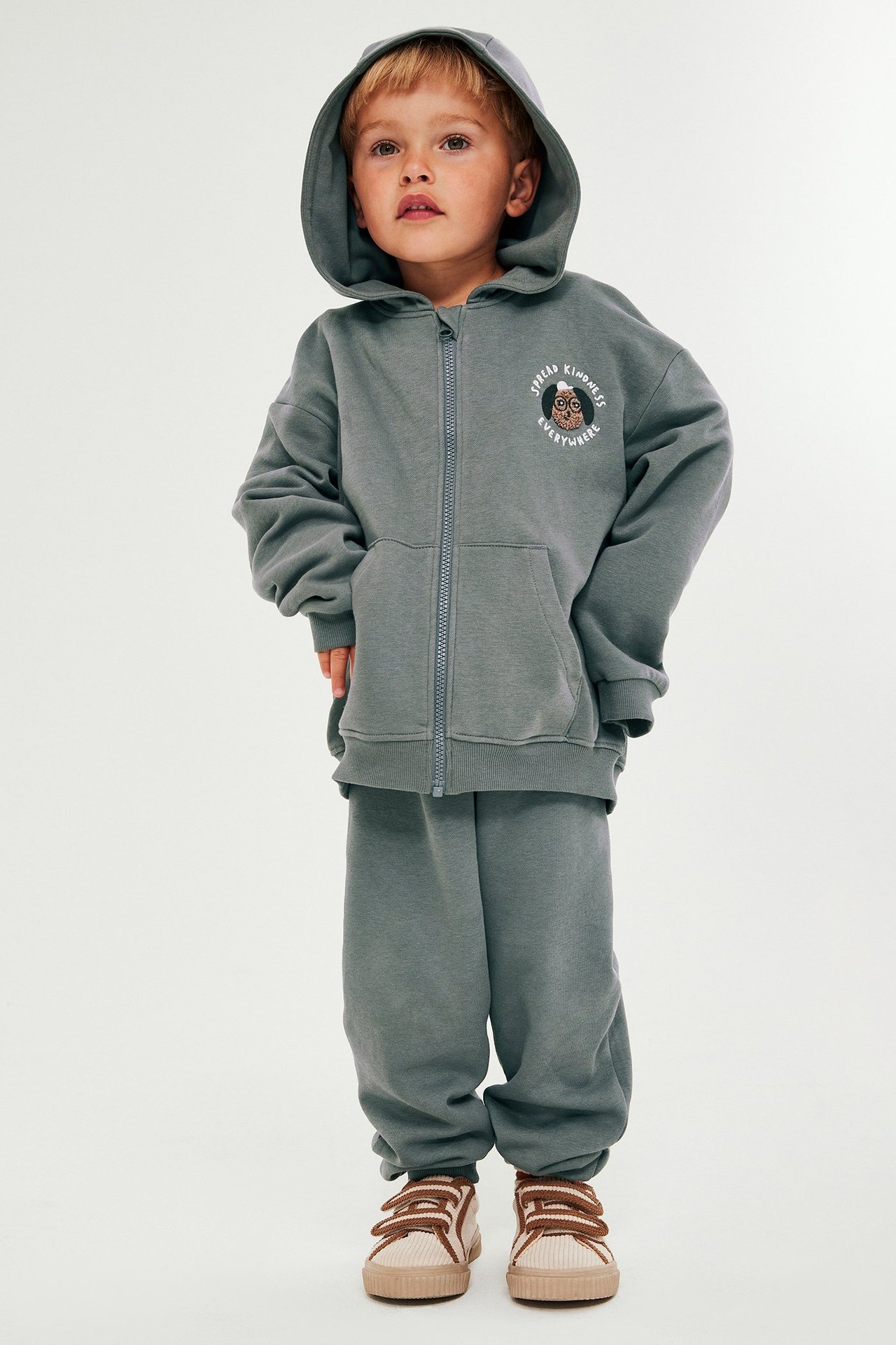 Edestä: Lapsi harmaassa hupullisessa vetoketjutakissa ja collegehousuissa, beiget tennarit jalassa.