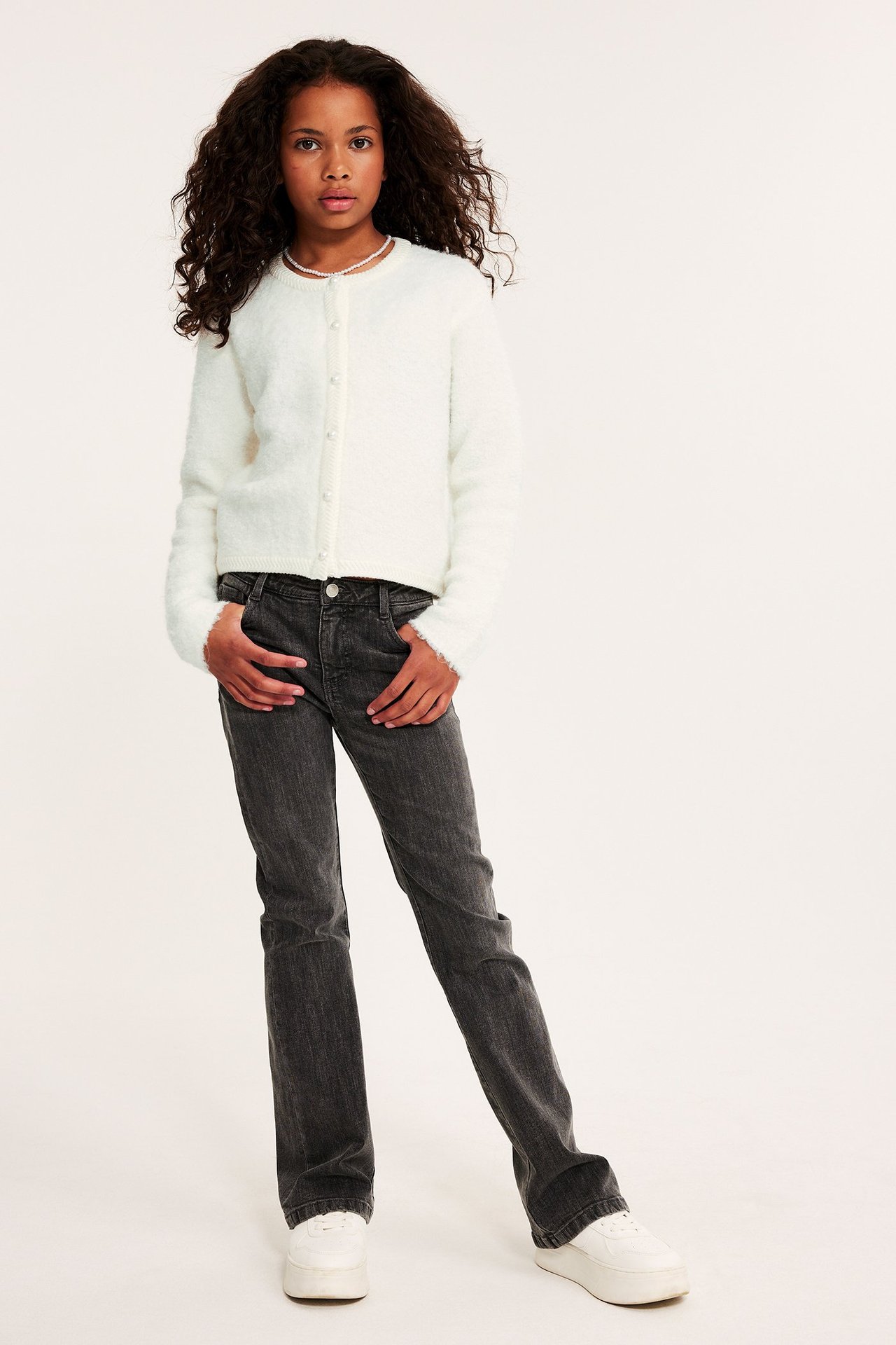 Edestä: Lapsi luonnonvalkoisessa neuletakissa helmipainikkeilla, helmikaulakoru, tummanharmaat farkut ja valkoiset lenkkarit.