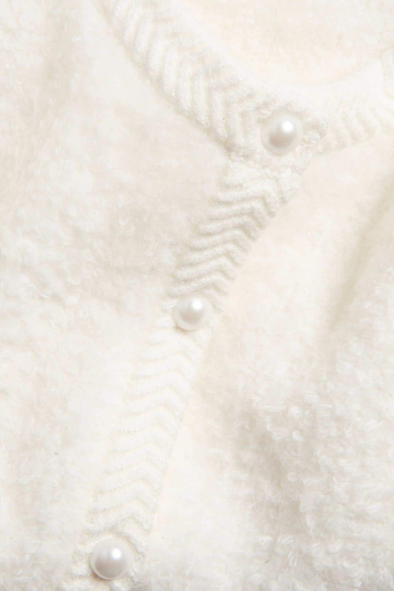 Zbliżenie na kremowy dzianinowy kardigan z perłowymi guzikami i zygzakowatą listwą.