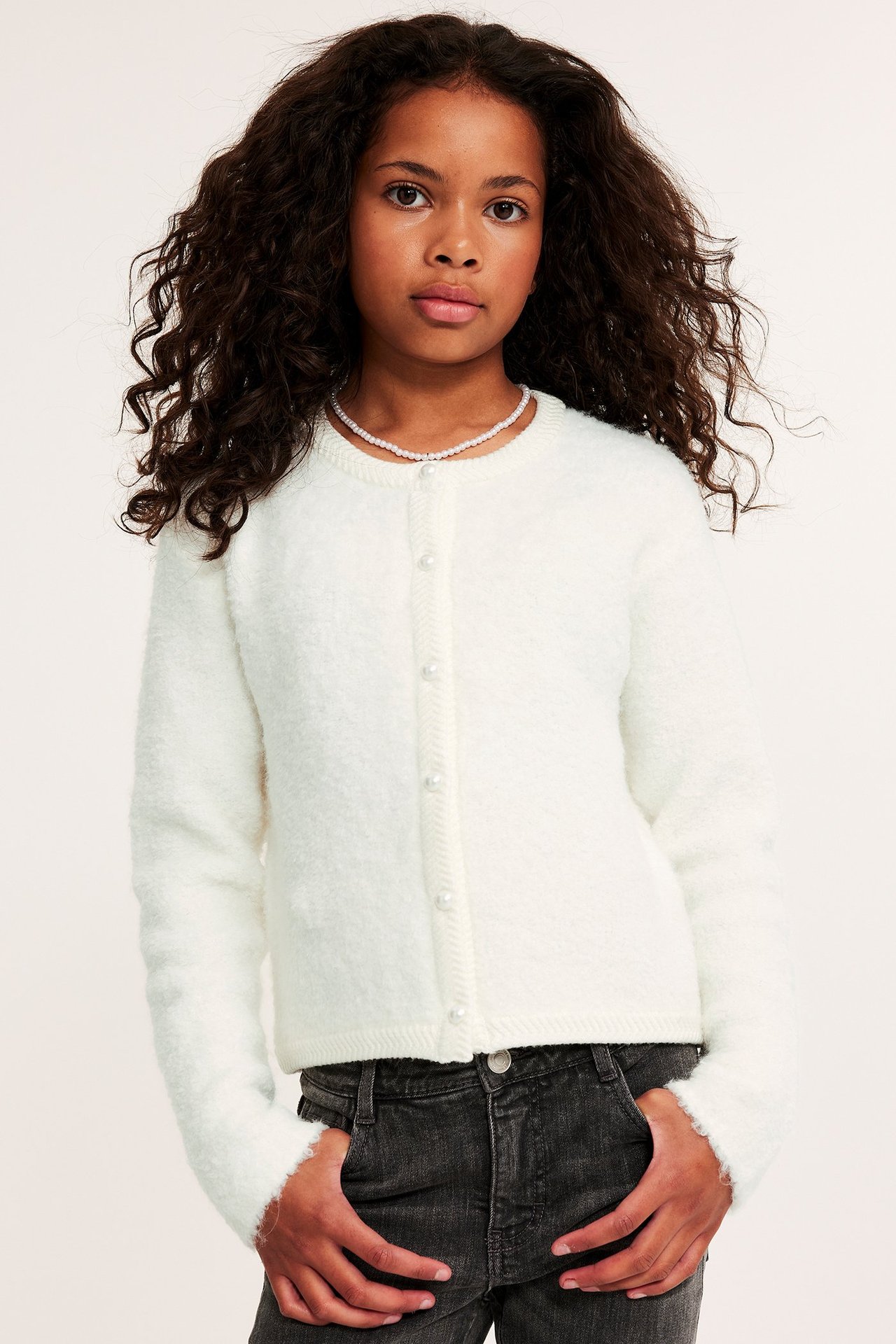 Widok z przodu: Dziecko w kremowym swetrze rozpinanym z perłowymi guzikami, naszyjniku i ciemnych dżinsach.
