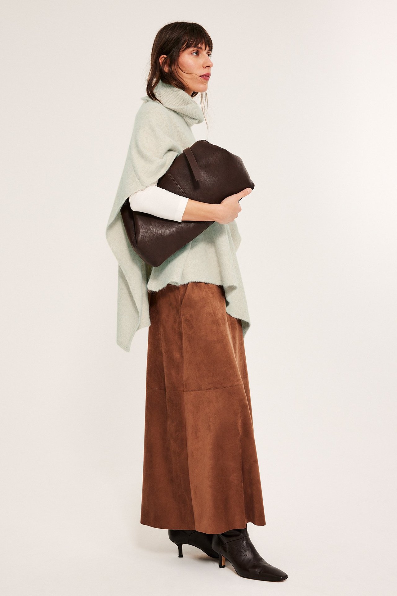 Kvinne i beige melert poncho med polo, hviler arm på svart støvlet.