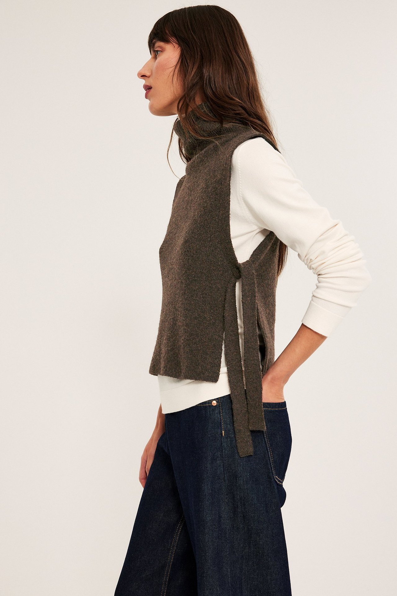 Kvinna bär en brun melange stickad väst med krage och knytband, åtsittande över en vit tröja.