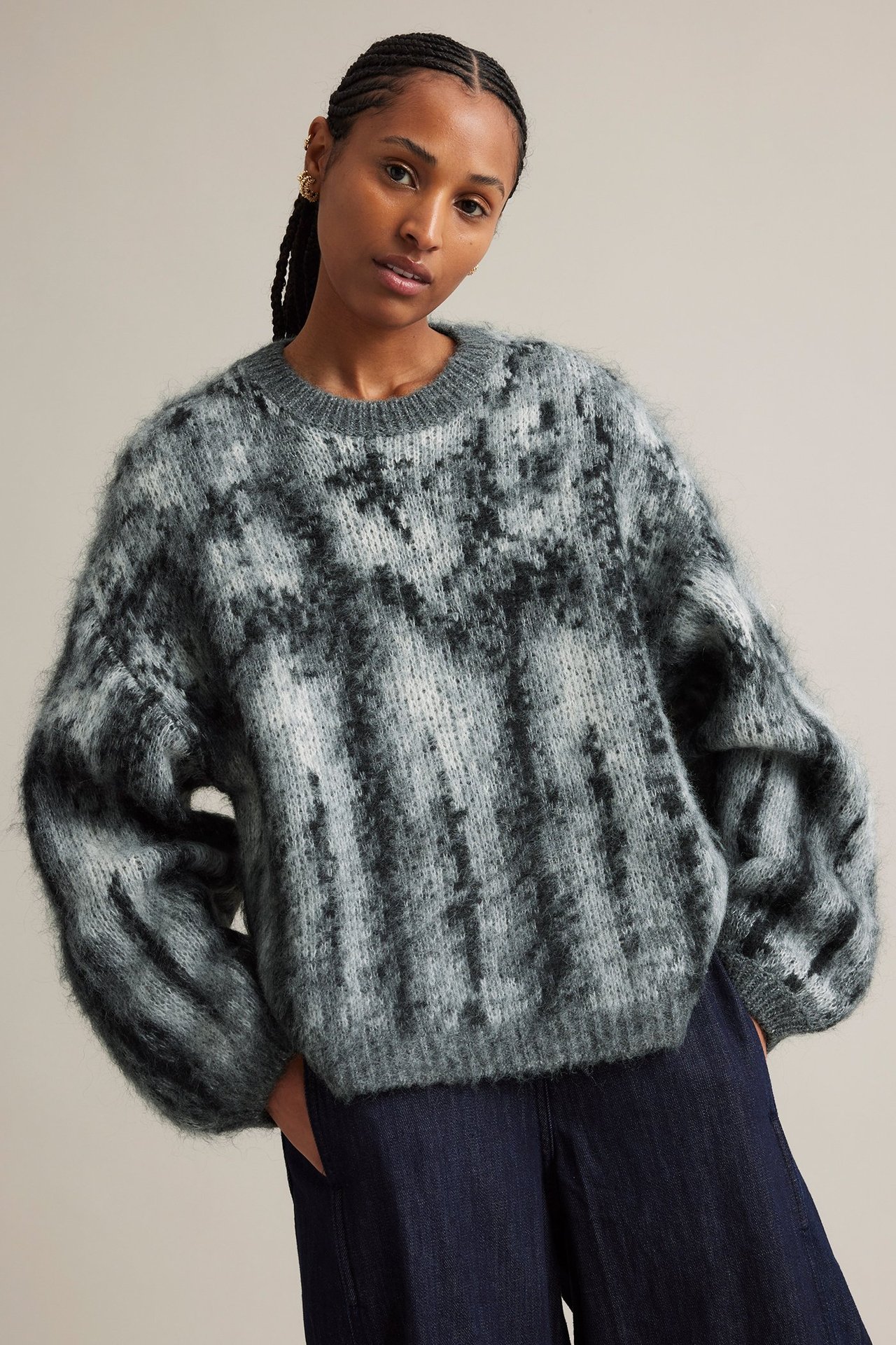 Framsida: Kvinna i fluffig svart och grå jacquardstickad tröja med vida ärmar och mörkblå byxor.