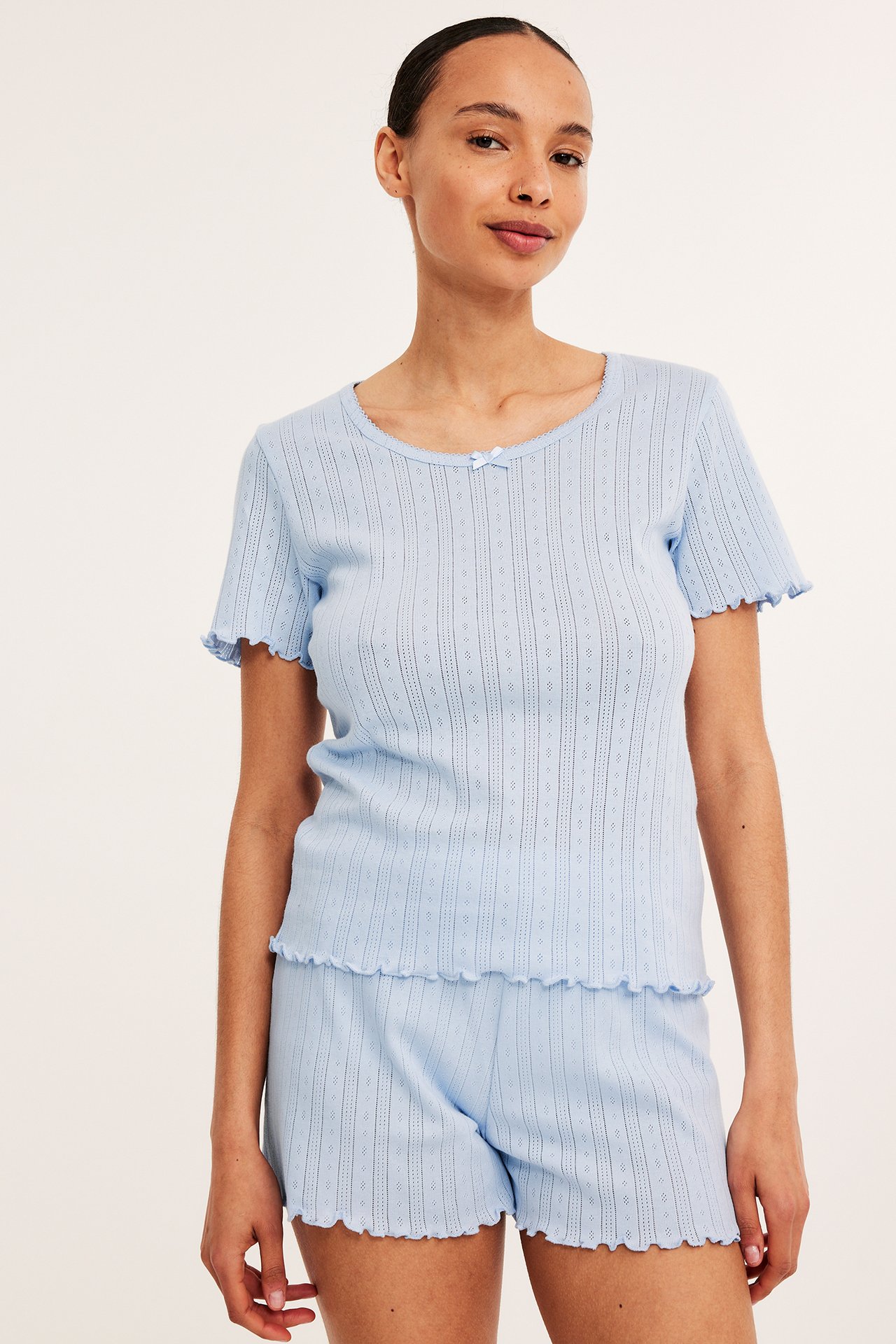 Frontbilde av kvinne i lyseblått pointelle-strikket pyjamassett med volangkanter.