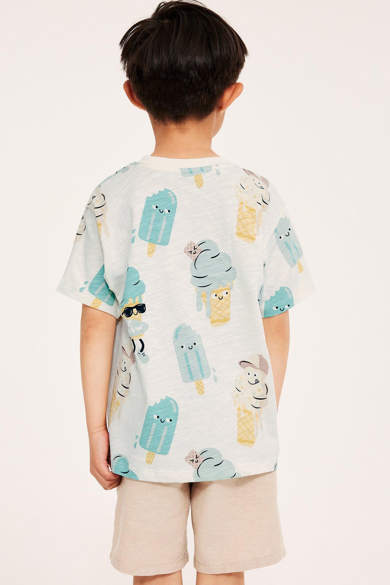 Widok z tyłu dziecka w kremowym, oversize'owym bawełnianym T-shircie z nadrukiem lodów i beżowych szortach.