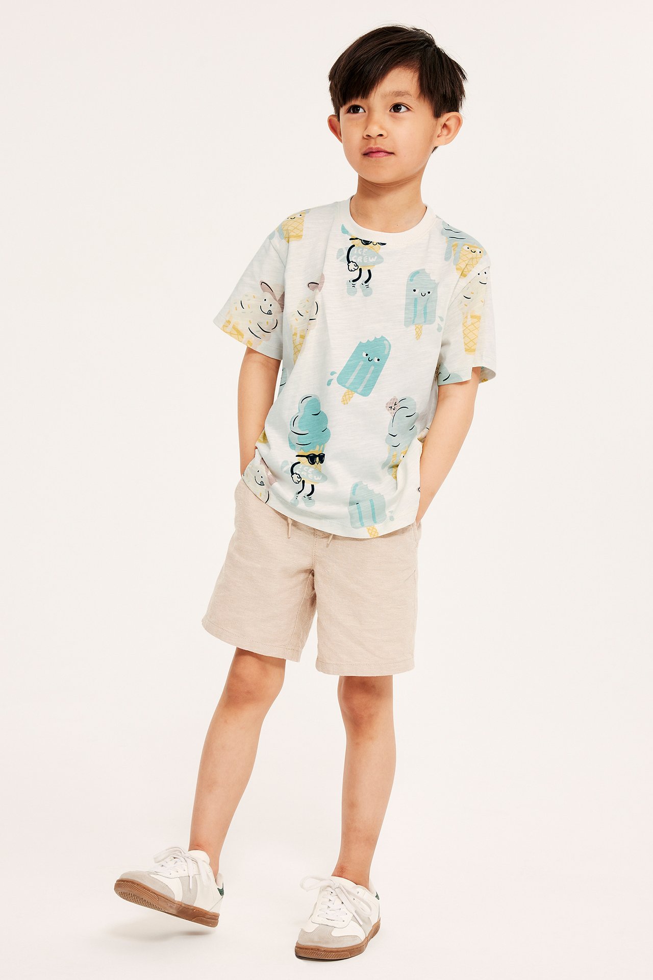Dziecko w kremowym T-shircie oversize z nadrukiem lodów, beżowe szorty, białe sneakersy.
