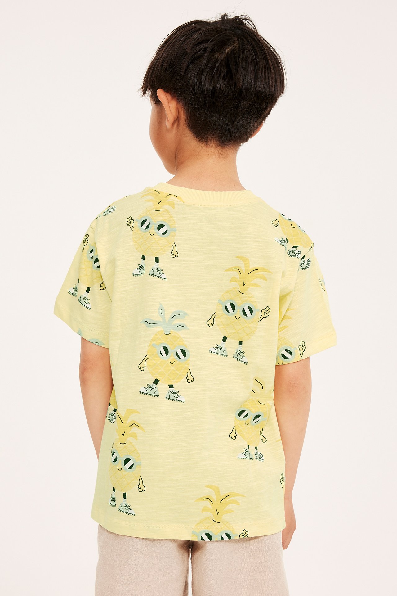 Widok z tyłu dziecka w jasnożółtym bawełnianym T-shircie oversize z nadrukiem ananasów i jasnobeżowych szortach.