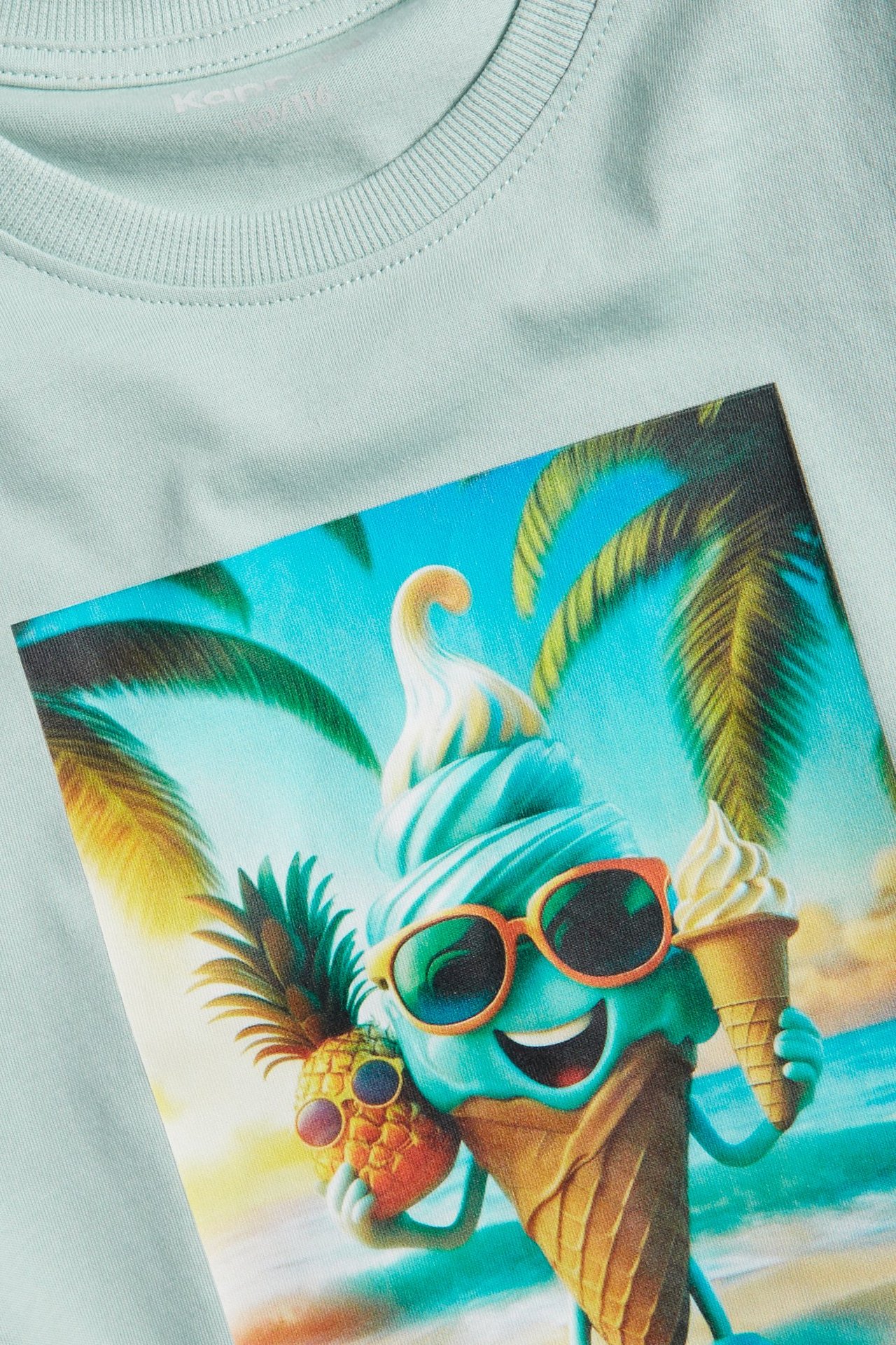 Zbliżenie na jasnoturkusową bawełnianą koszulkę dziecięcą z nadrukiem lodów i ananasa.