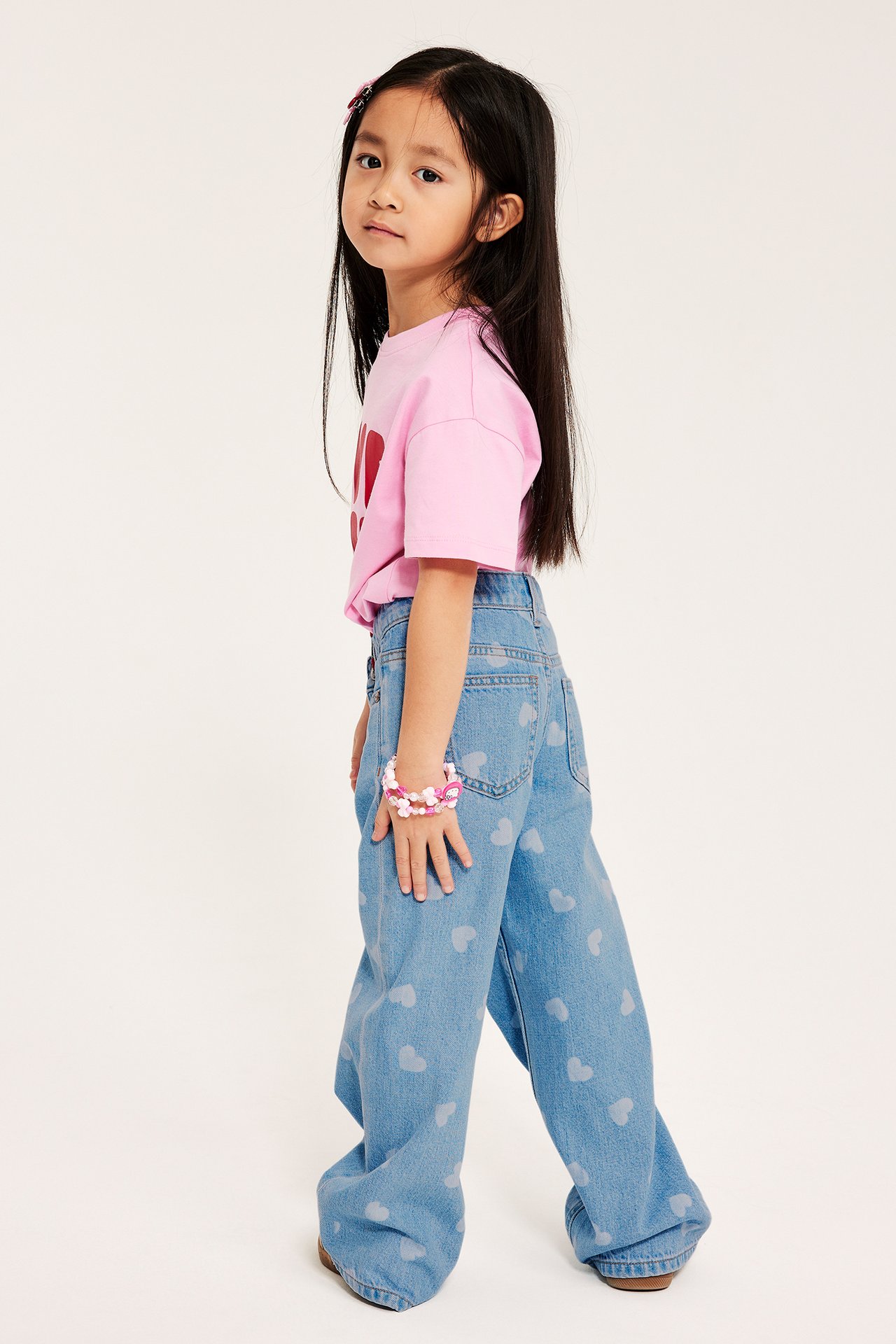 Sida vy av ett barn som bär ljus denim vida jeans med hjärtmönster, en rosa t-shirt och ett armband.