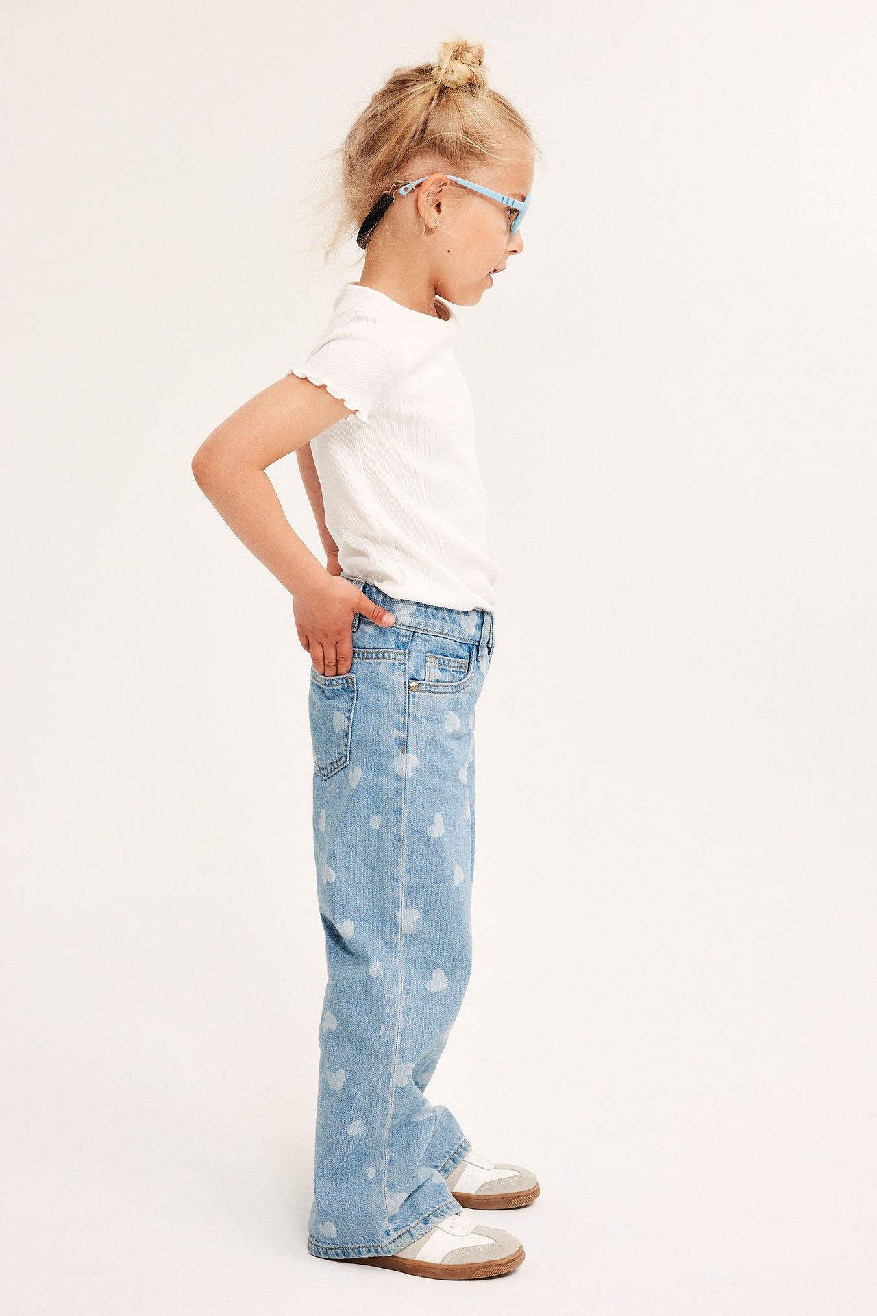 Sidovy: Barn i ljusblå vida jeans med vita hjärtan, vit t-shirt, blå glasögon, sneakers.