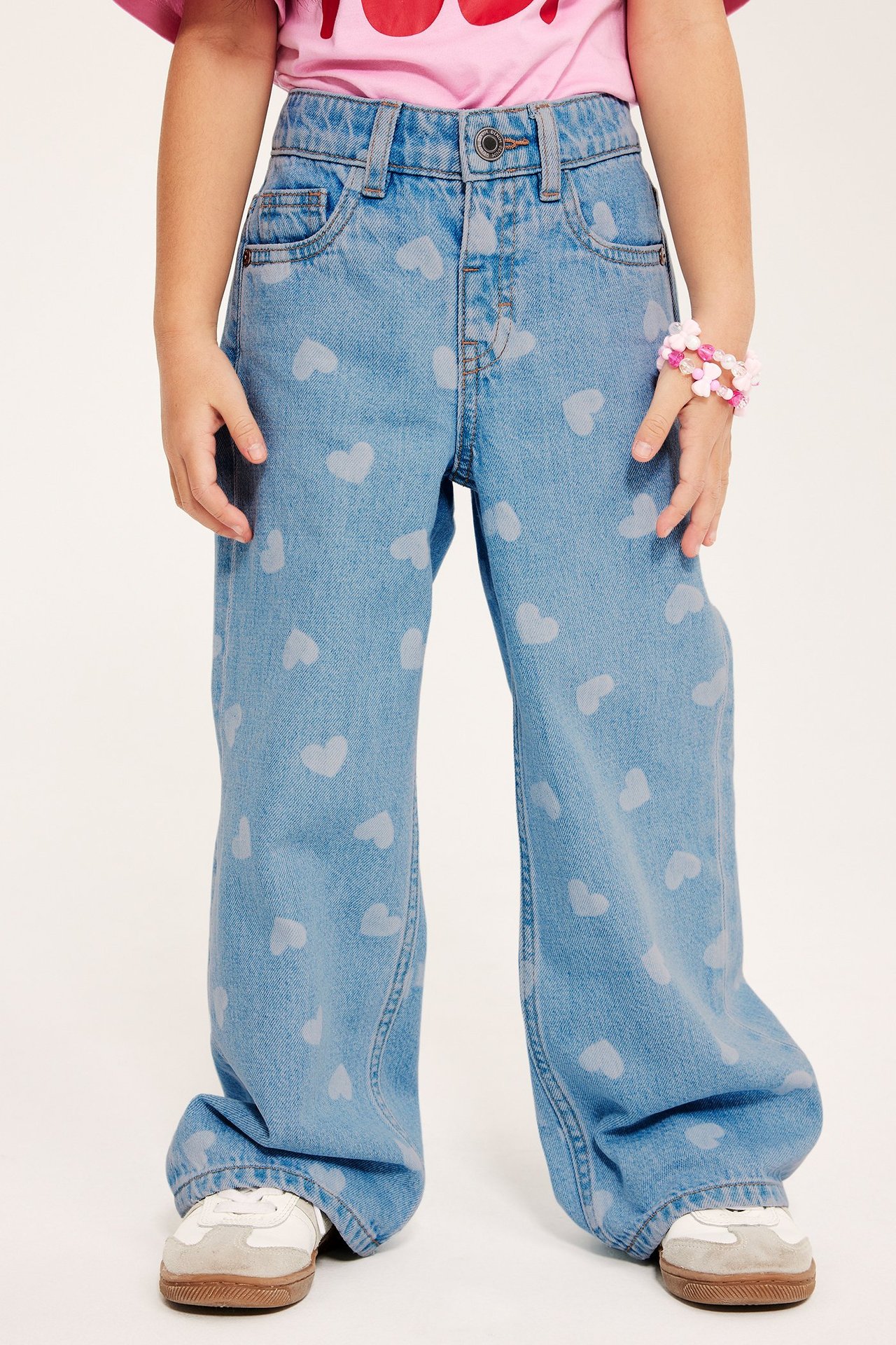 Barn i lyseblå vide denimjeans med hjerteprint, rosa topp, hvite joggesko og rosa armbånd, frontvisning.