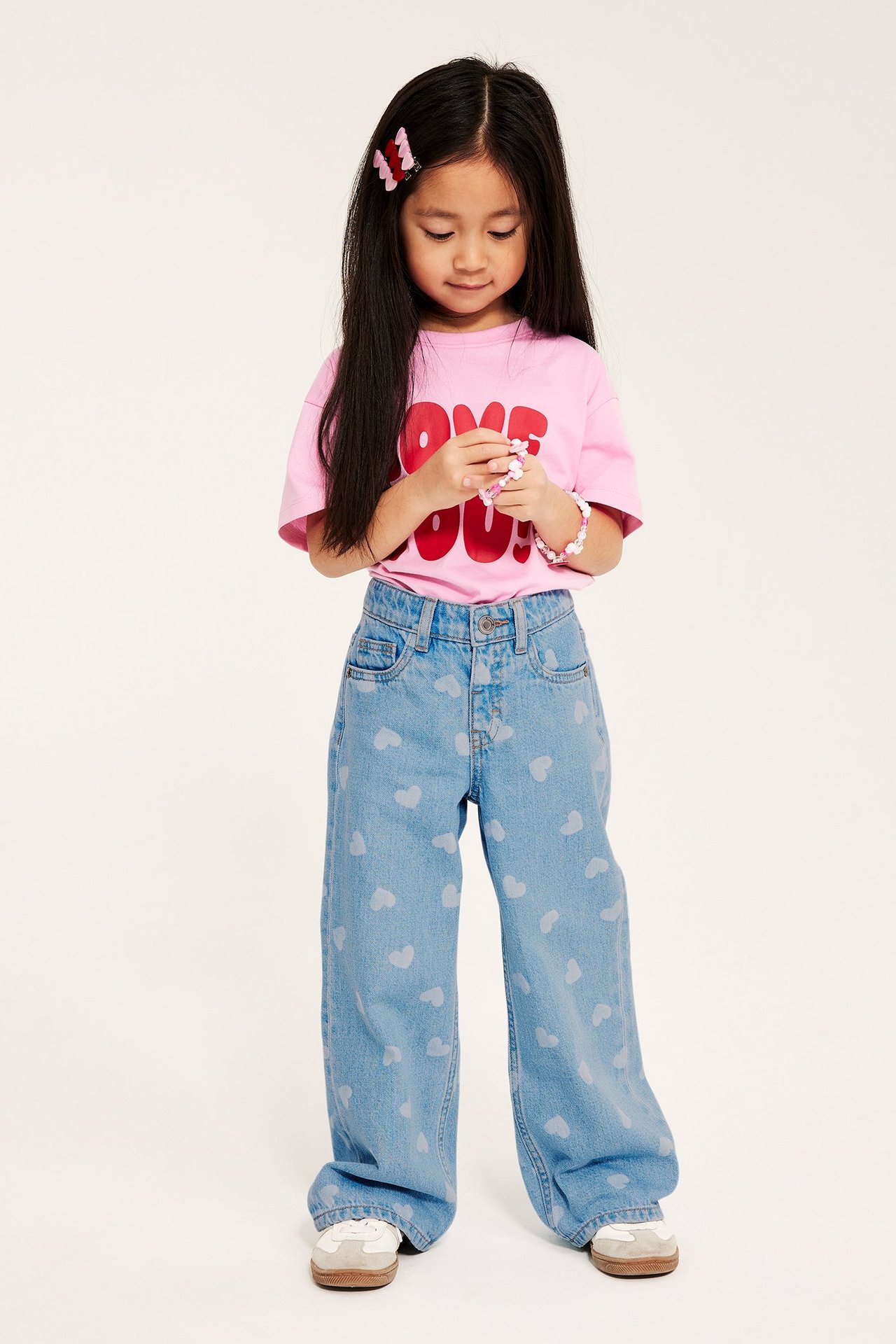 Etunäkymä: Lapsi vaaleansinisissä sydänkuvioisissa leveissä farkuissa, vaaleanpunaisessa t-paidassa, hiusklipseillä, pitäen ranneketta.