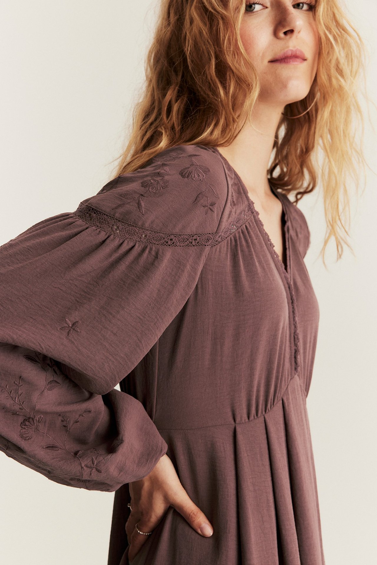 Lähikuva naisesta yllään viininpunainen brodeerattu polyesterimekko pitkillä pussihihoilla ja pitsiyksityiskohdilla.