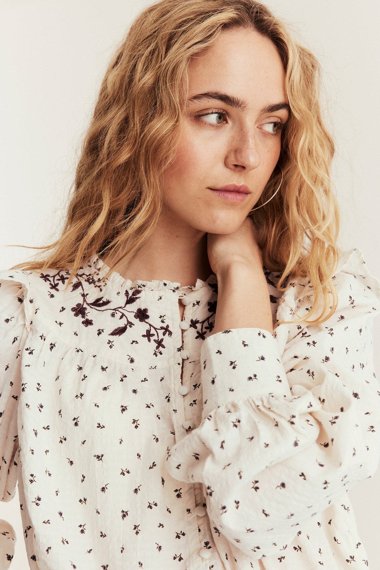 Kvinna i ljusbeige blus med blommigt mönster, broderat ok, volanghals, puffärmar och ringörhängen.