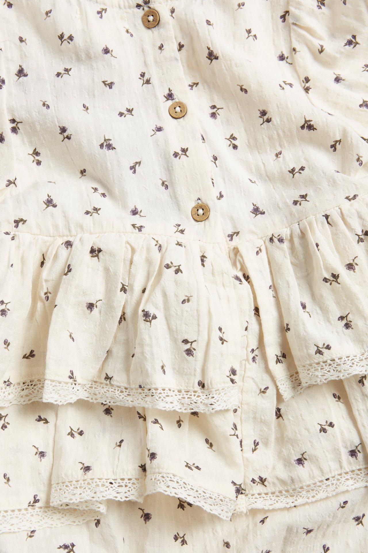 Zbliżenie na jasnobeżowe body niemowlęce w kwiaty, z falbankami, koronkowym wykończeniem i drewnianymi guzikami.