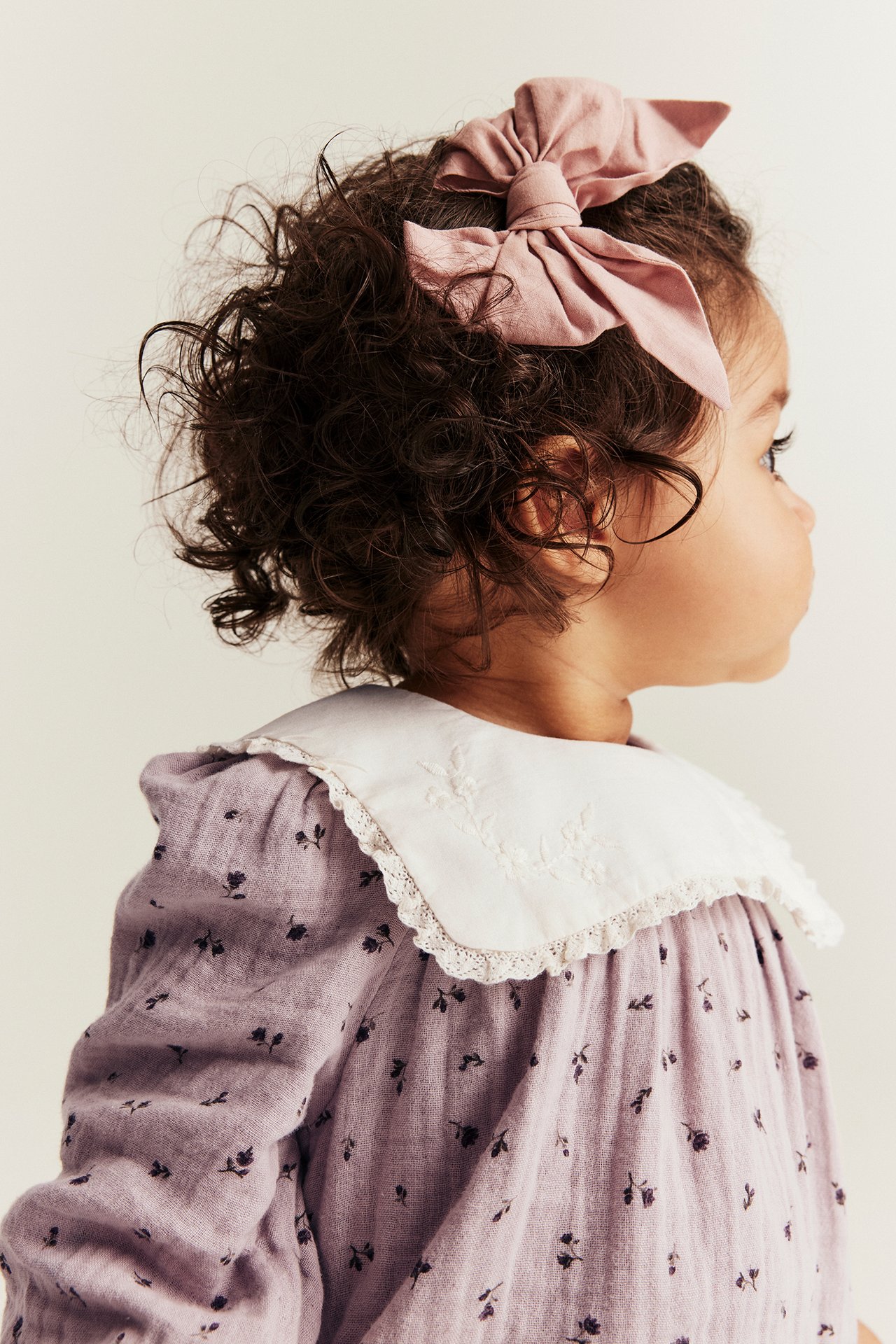 Sivukuva, vauva vaaleanlila kukkabody, valkoinen kirjailtu pitsikaulus ja vaaleanpunainen hiusrusetti.