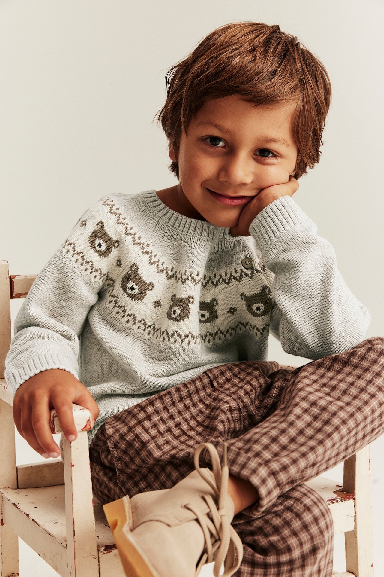 Edestä hymyilevä lapsi yllään beigemelattu nallekuvioinen neule ja ruskeat ruudulliset housut.