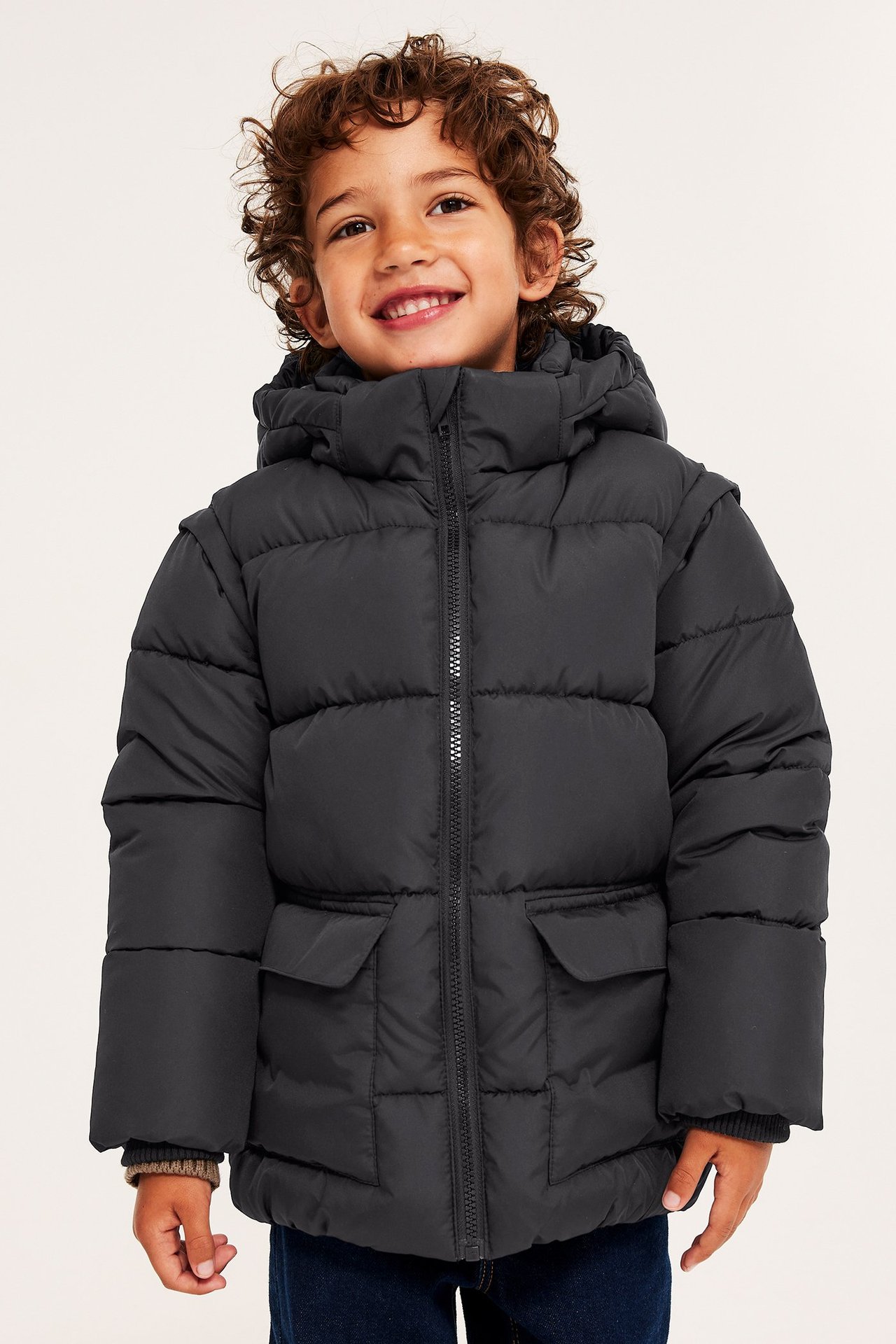 Frontbilde av et smilende barn i svart vattert boblejakke med hette og mørkeblå jeans.