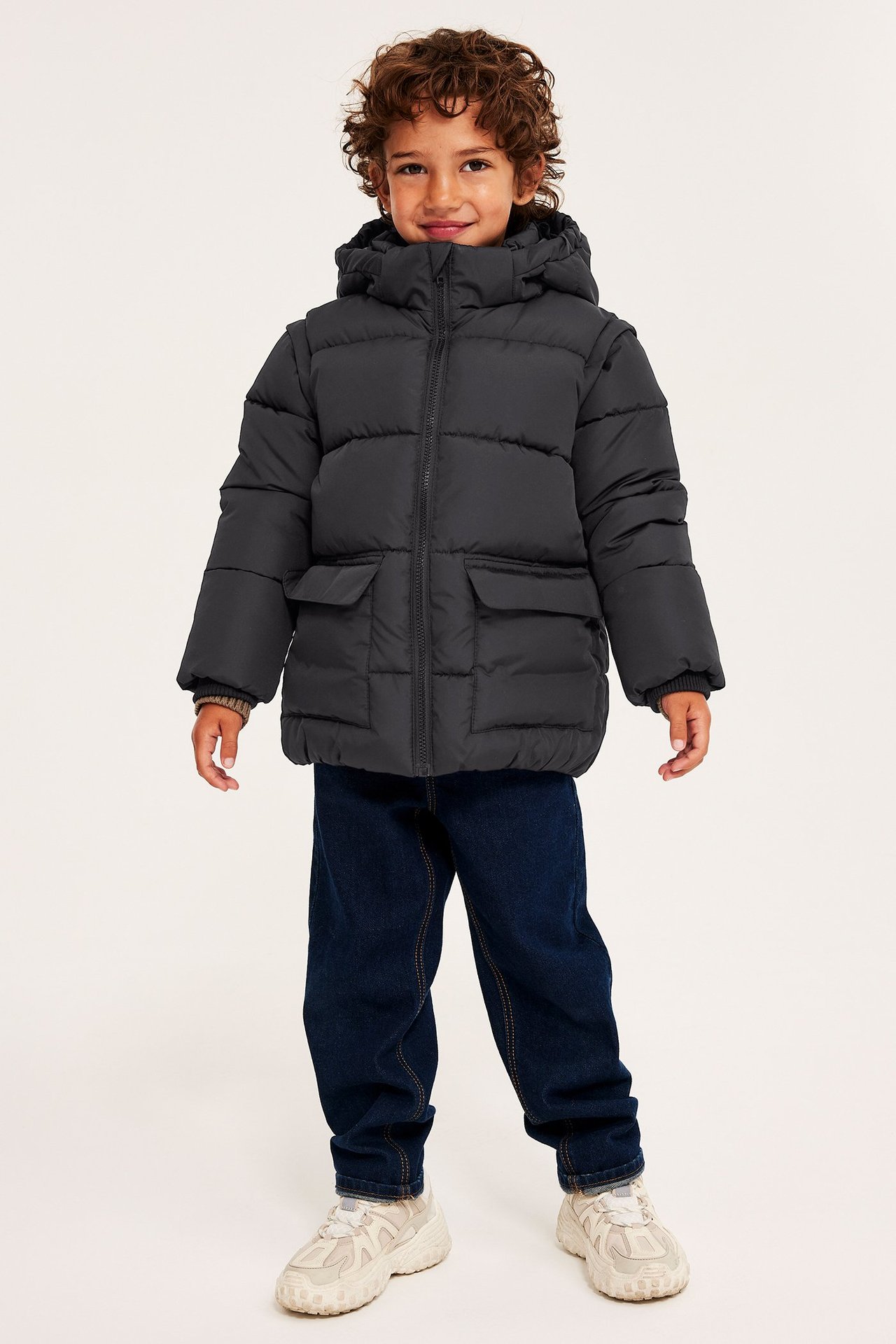 Forfra: Barn i svart vattert pufferjakke med hette, mørkeblå jeans og lyse joggesko.