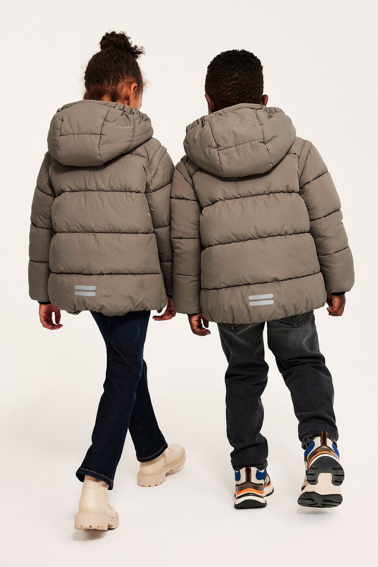 Bakfra: to barn i brune, hette-pufferjakker med refleksstriper, jeans og sko.