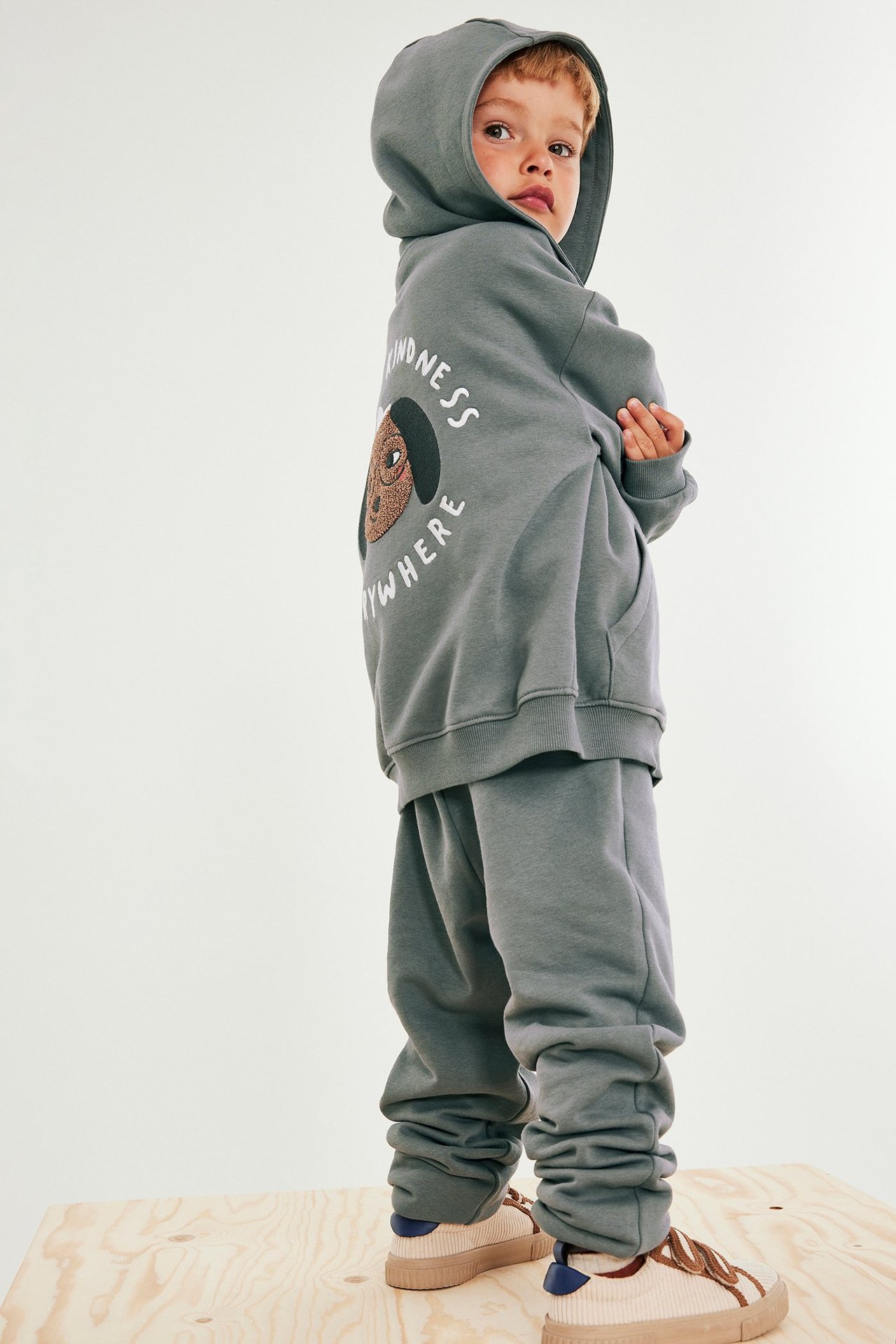 Sidovy: barn i grå hoodie med hundbrodyr på ryggen, matchande mjukisbyxor och beiga sneakers.