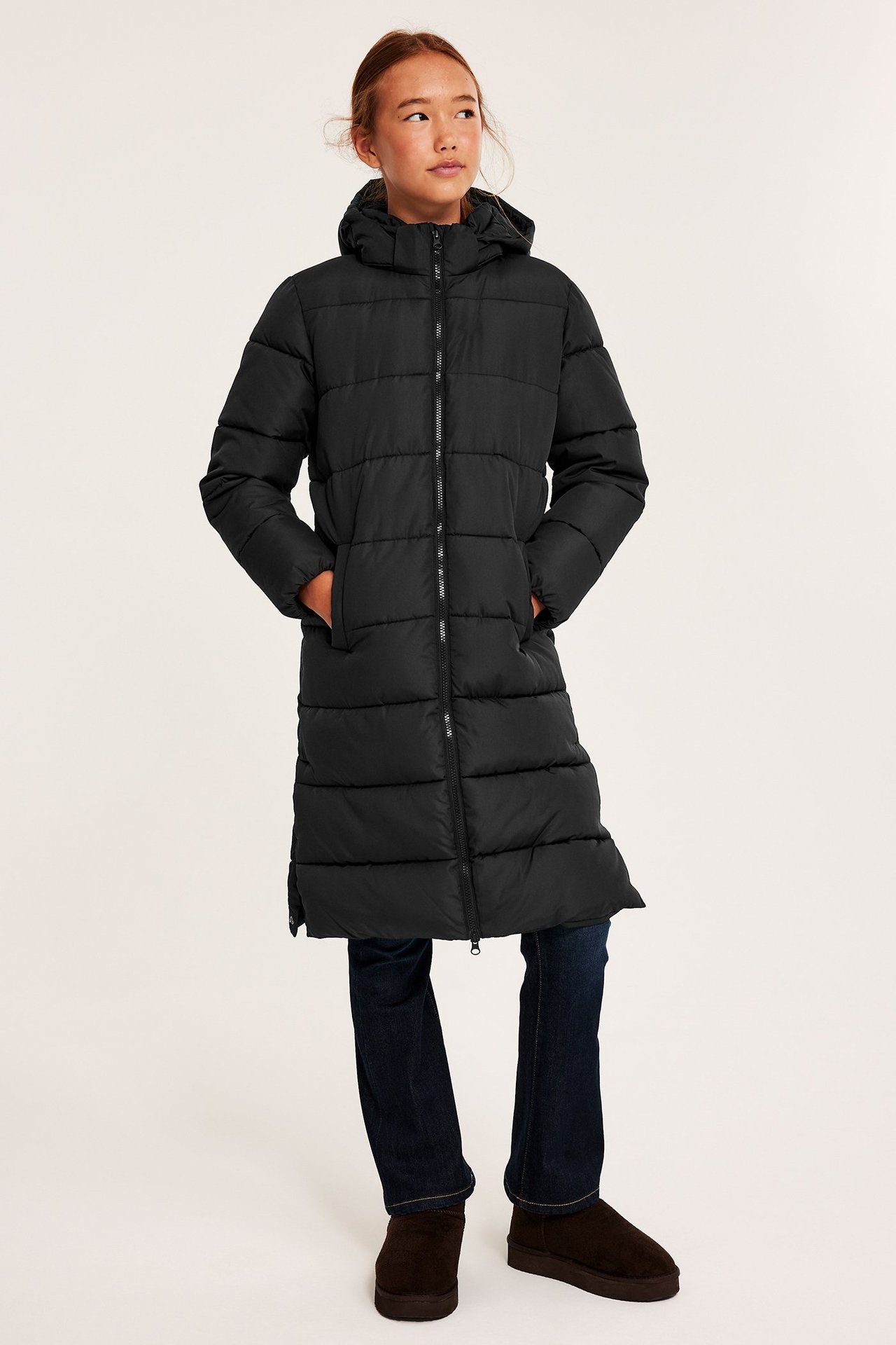 Framifrån: Barn i lång svart pufferjacka med huva, mörkblå jeans och bruna ankelboots.