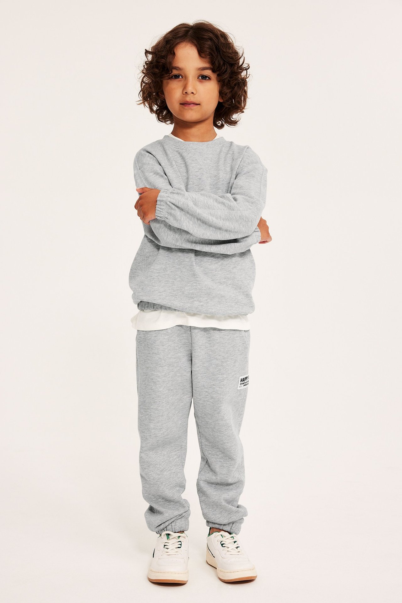 Widok z przodu: Dziecko w szarych melanżowych spodniach dresowych, pasującej bluzie, białym t-shircie i białych sneakersach.