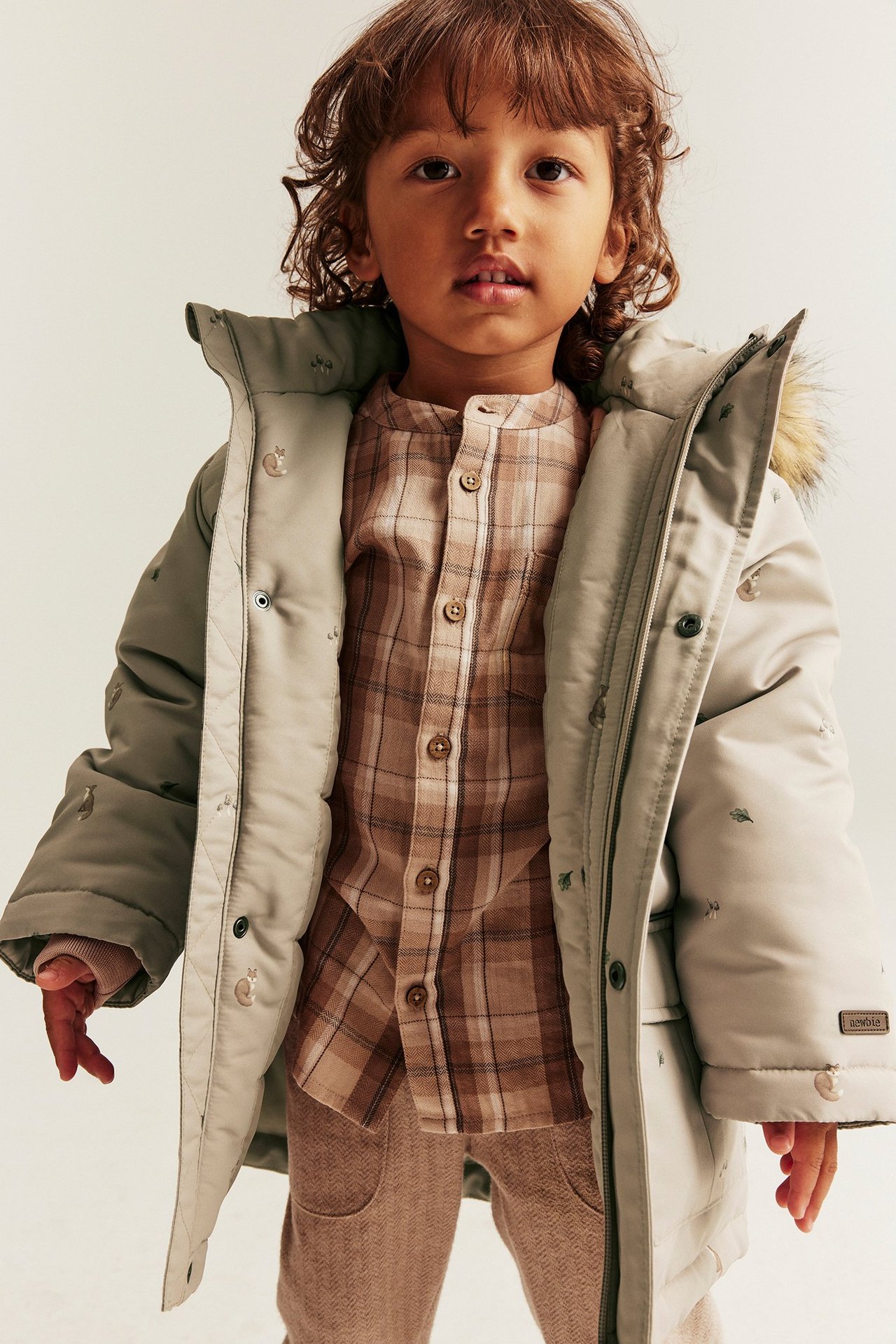 Frontvy på ett barn i öppen beige vadderad jacka med rävmönster över rutig skjorta och beige byxor.
