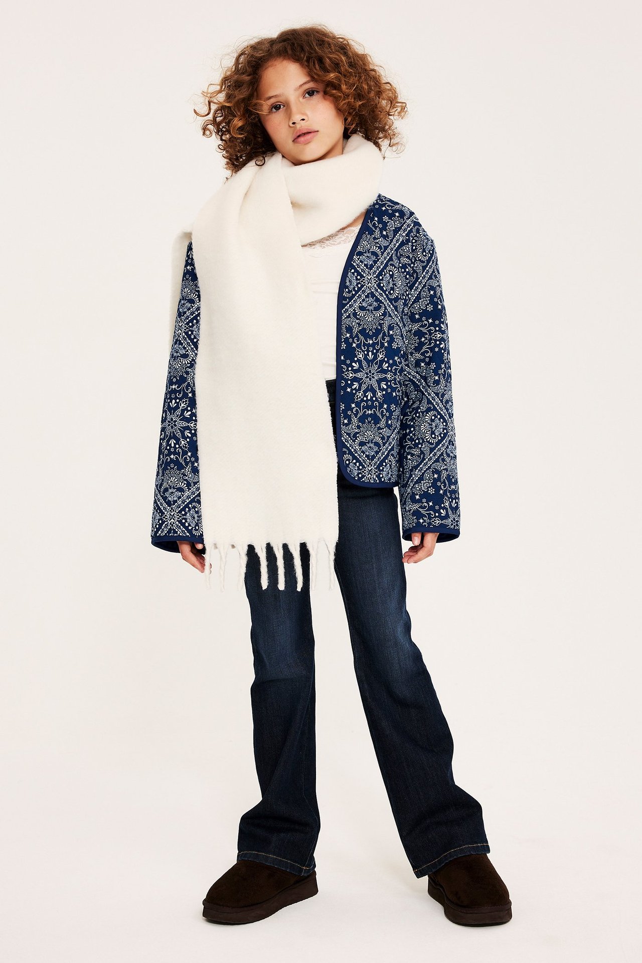 Framifrån: Kid i blå mönstrad quiltad jacka, gräddvit halsduk, vit topp, mörkblå jeans, bruna skor.