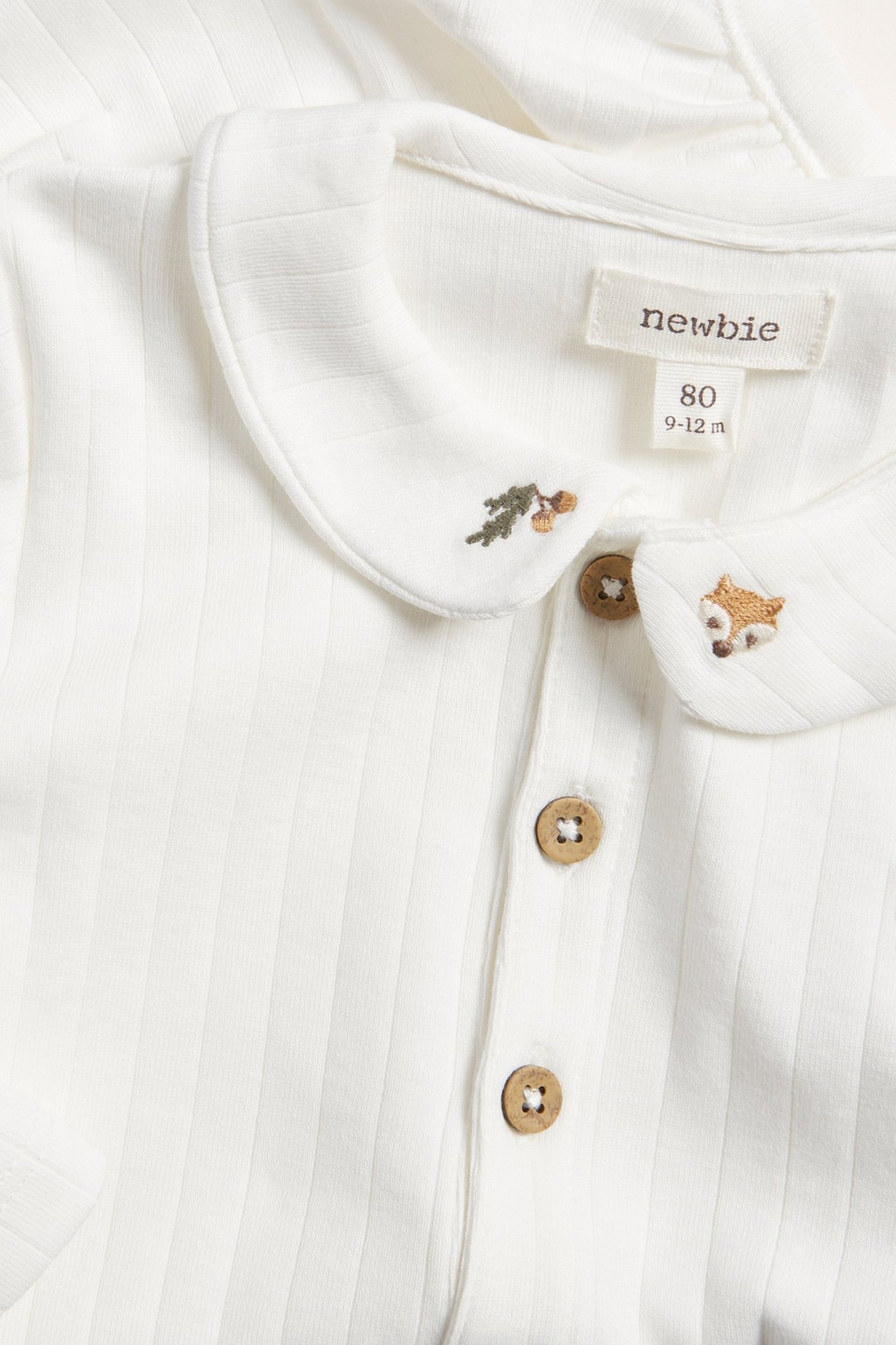 Lähikuva luonnonvalkoisesta ribatusta vauvan body-paidasta, jossa on pitkät hihat, kaulus ja kettu-kirjailu.
