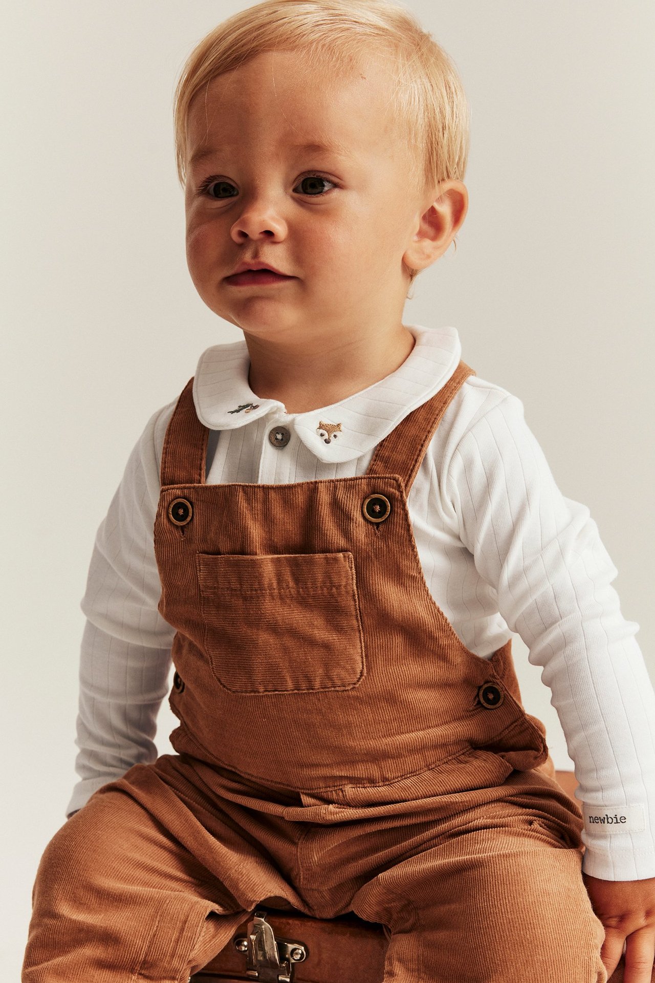 Lähikuva edestä vauvasta yllään luonnonvalkoinen ribbattu body kettukirjonnalla ja ruskeat vakosamettihaalarit.