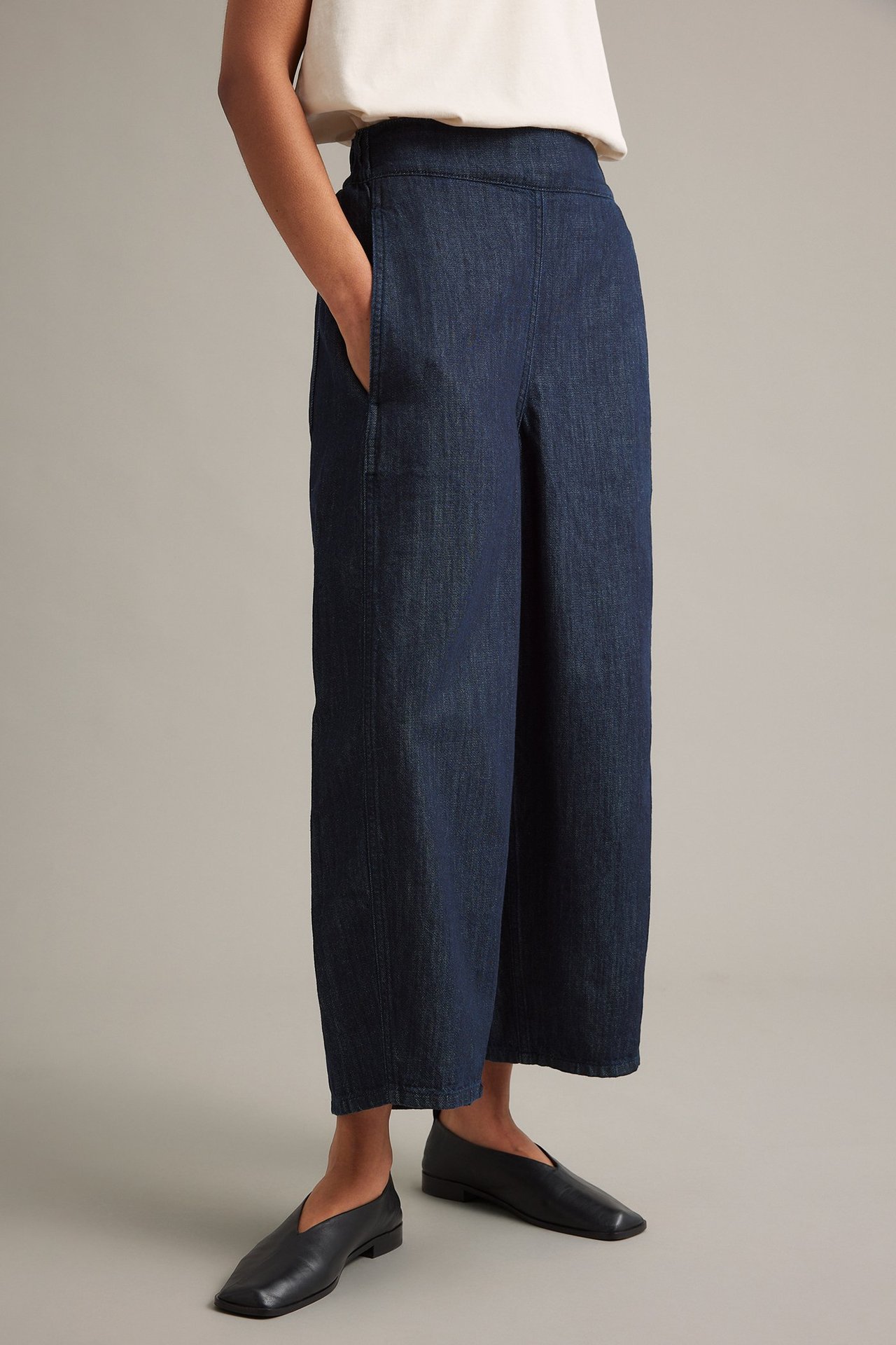 Frontvy av en kvinna i mörkblå barrel leg pull on-jeans, en ljus topp och svarta platta skor.