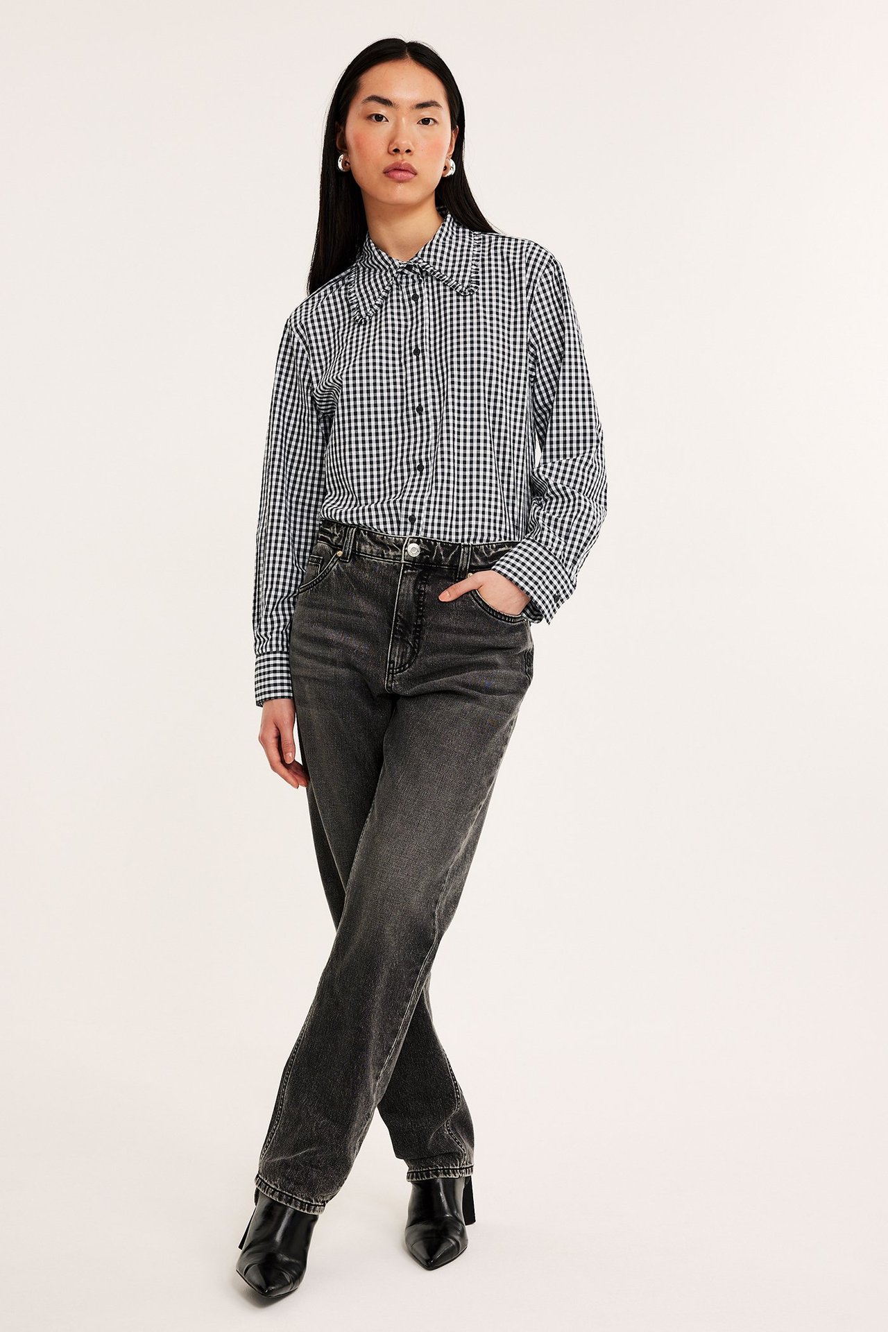 Frontvy: Kvinna i svartvit ginghamrutig bomullsskjorta med volangkrage, mörkgrå jeans och svarta stövlar.