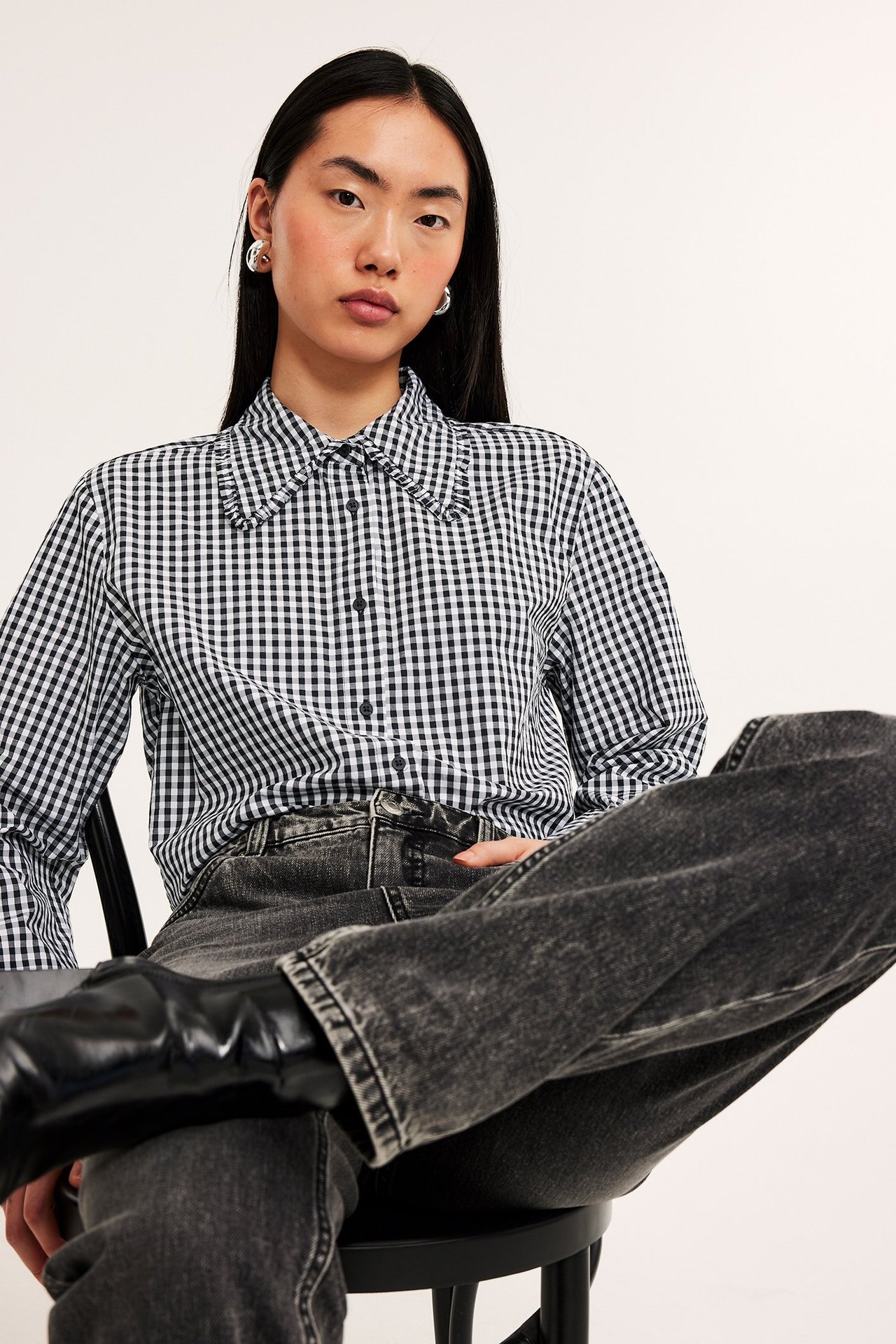 Widok z przodu: kobieta w czarno-białej koszuli bawełnianej w kratkę z falbanką, ciemnoszarych dżinsach, czarnych butach.