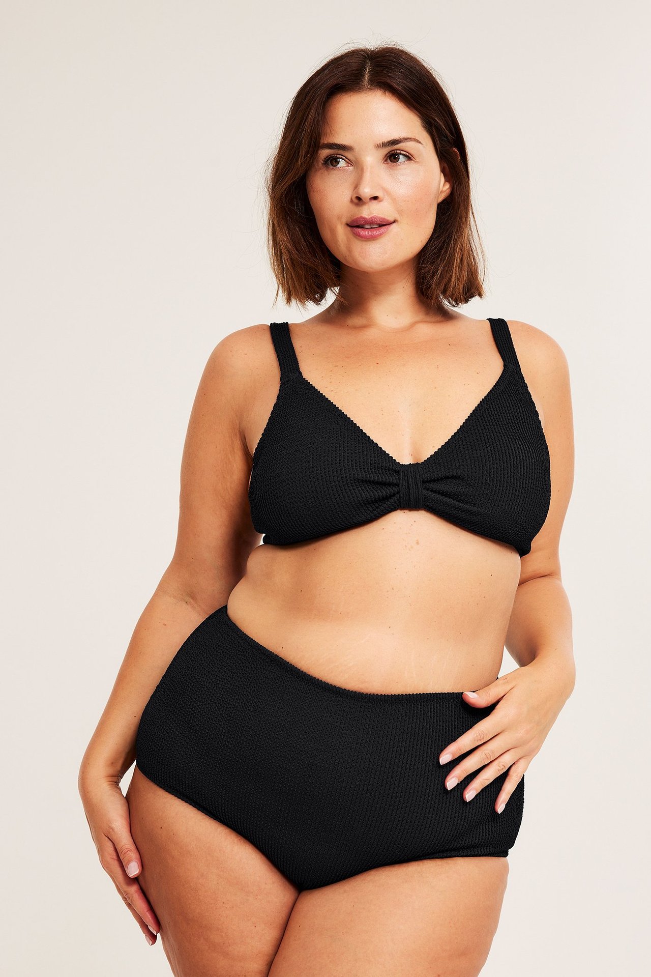 Przód: kobieta w czarnych, teksturowanych, wysokich majtkach kąpielowych i pasującej górze bikini.