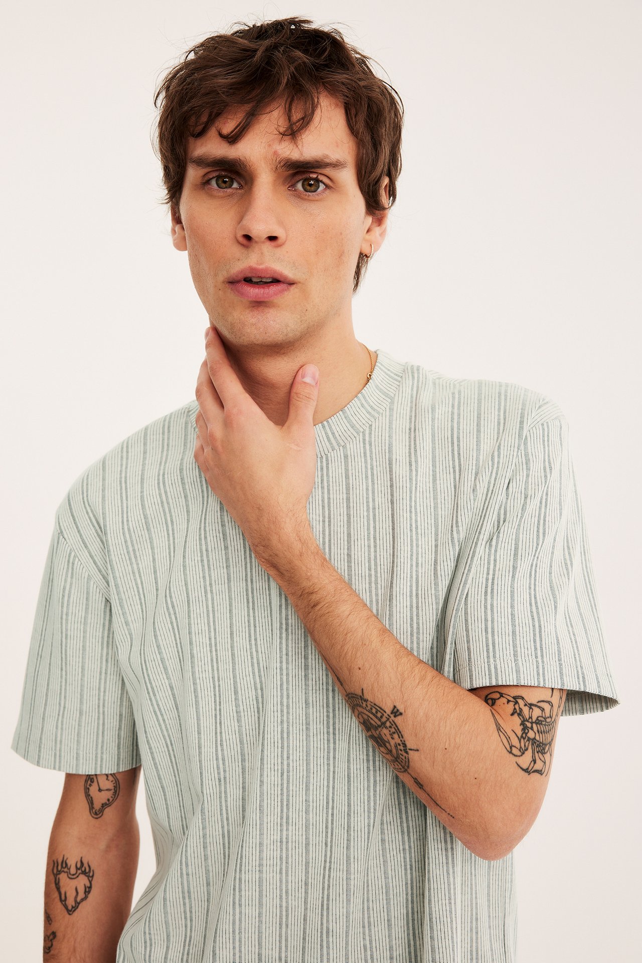 Lähikuva edestä: mies yllään luonnonvalkoinen kuvioitu lyhythihainen t-paita, kultaketju ja korvakoru.