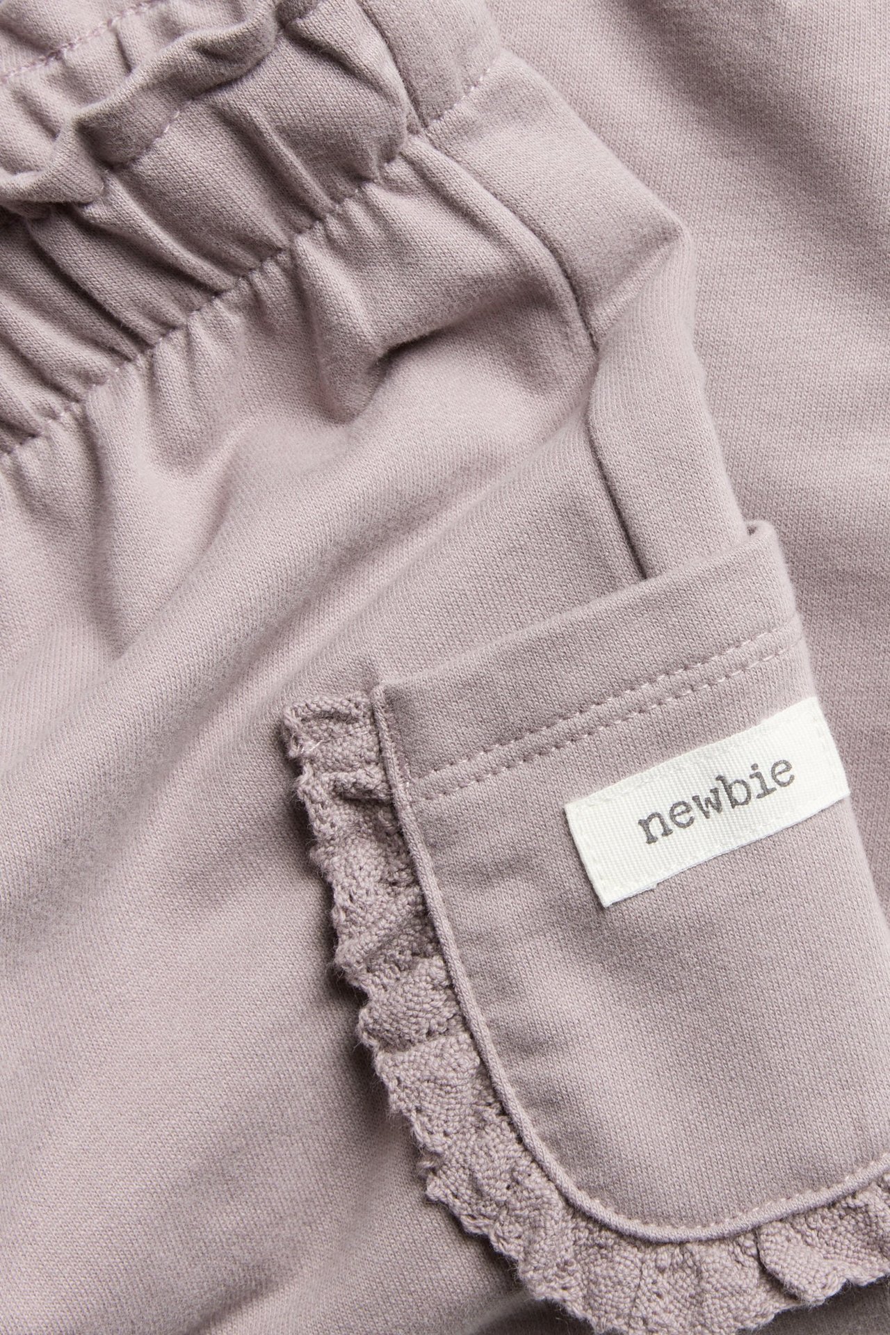Lähikuva vaaleanliiloista puuvillahousuista, joissa joustava vyötärö ja pitsireunainen tasku "newbie"-etiketillä.