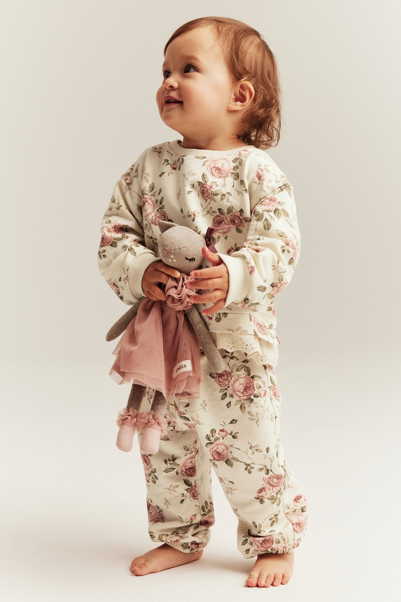 Forfra: Baby i lys beige blomstrete joggebukse med volangkant, matchende topp, holder en dukke.
