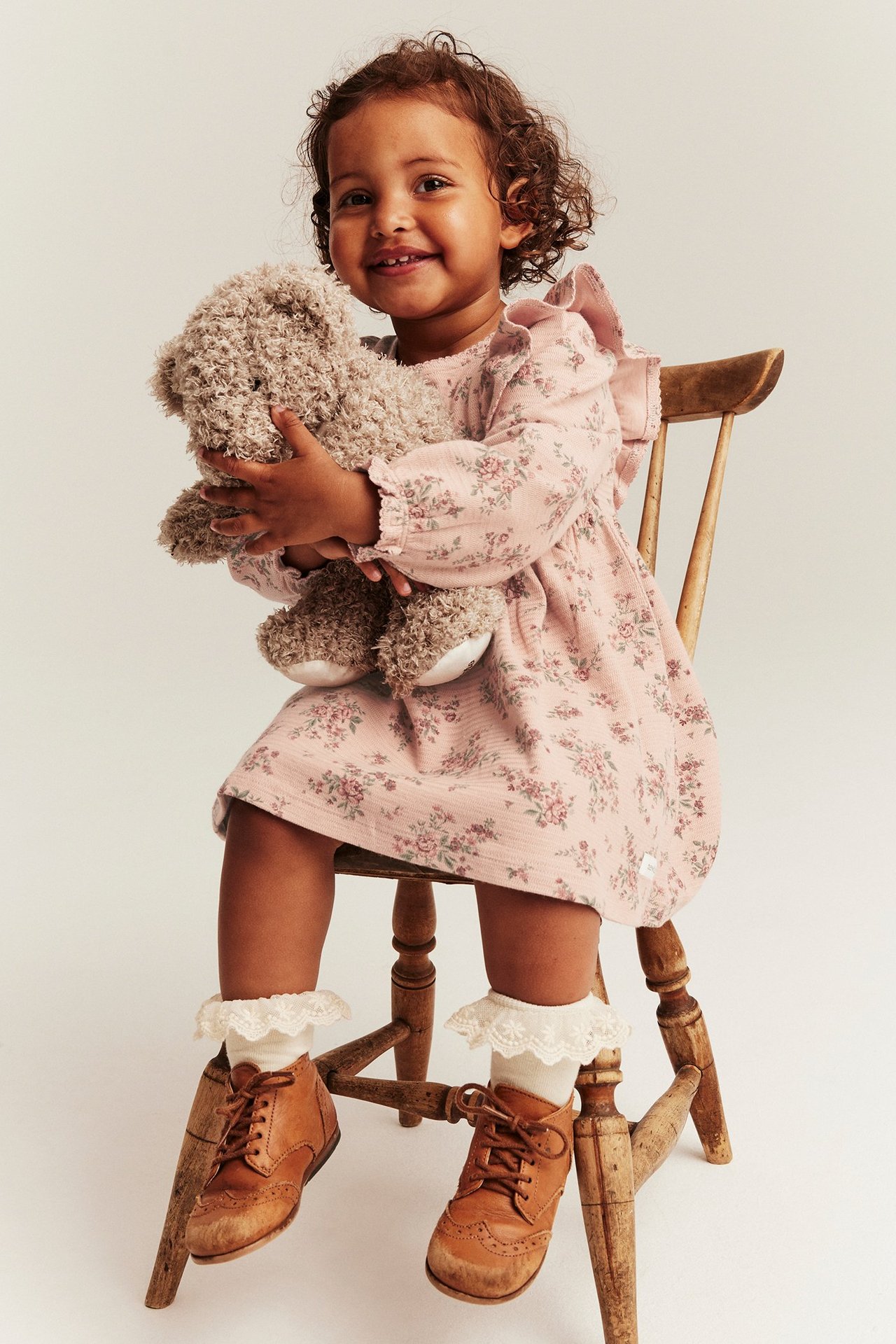 Barn i rosa blomstret jerseykjole, hvite blondesokker, brune sko, holder en teddybjørn.