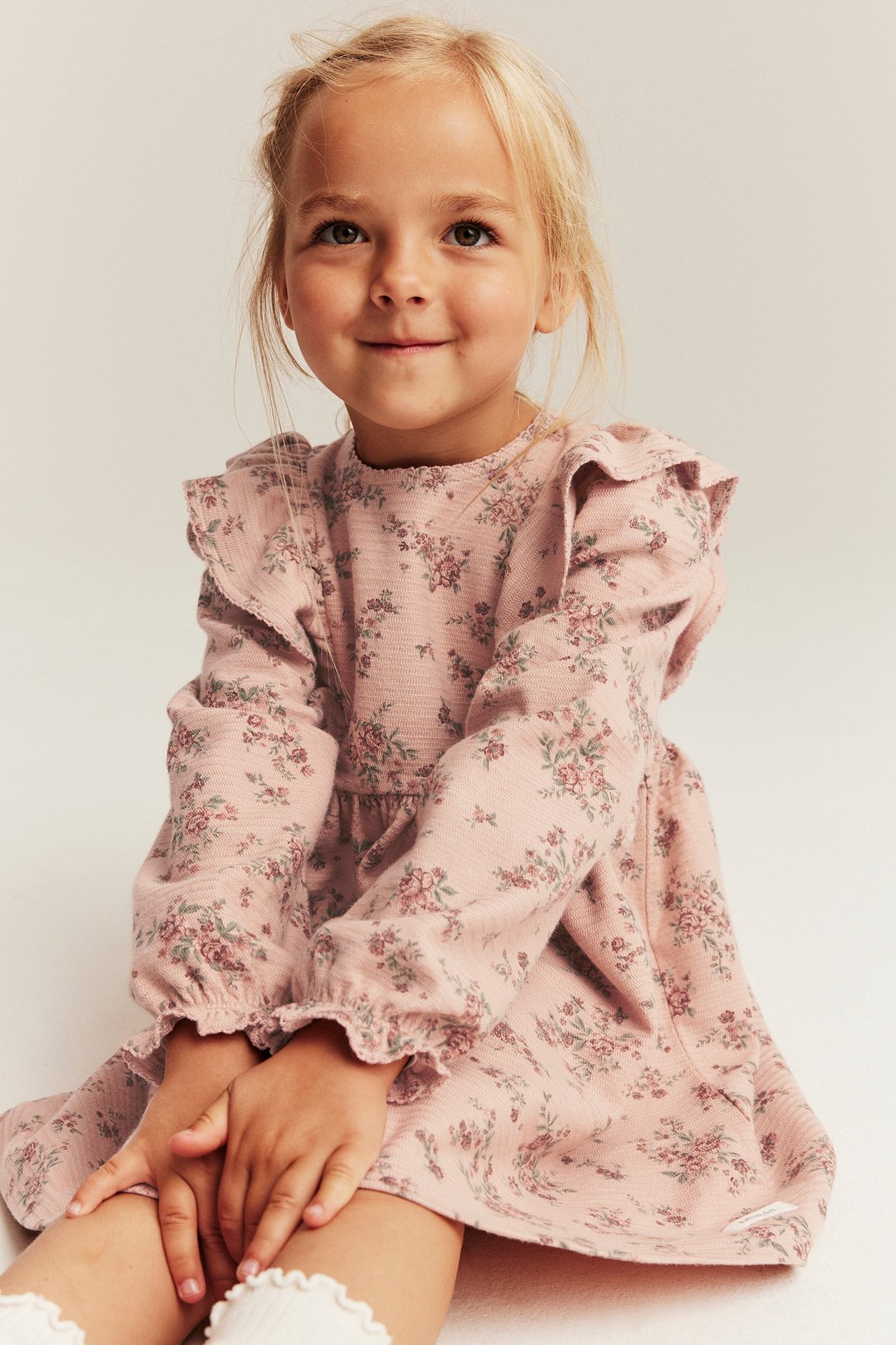 Forfra, et barn i rosa blomstret kjole i strukturjersey med volanger og hvite rysjesokker.