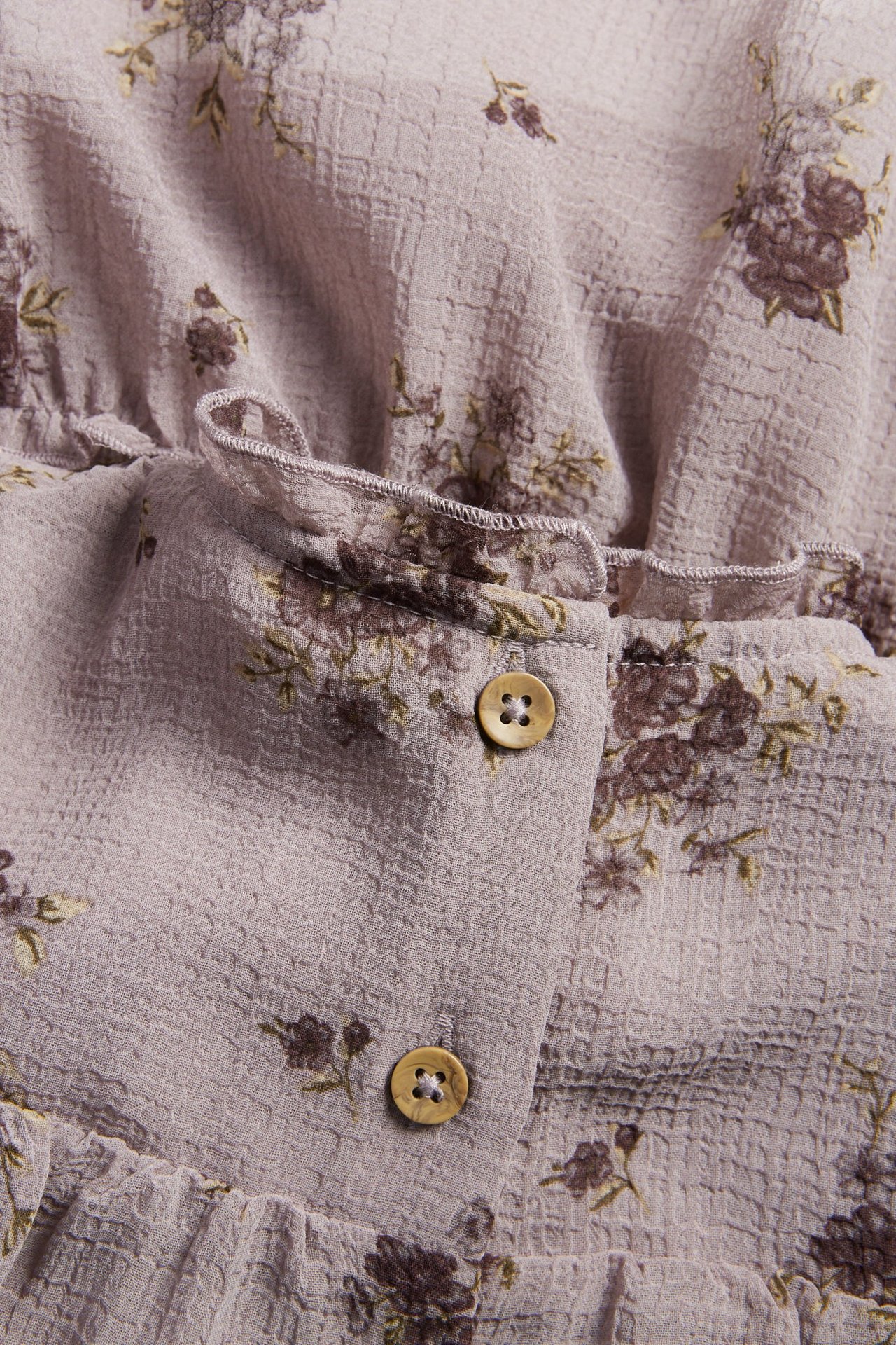 Zbliżenie jasnofioletowej kwiecistej szyfonowej sukienki z falbankami i dwoma jasnobrązowymi guzikami.