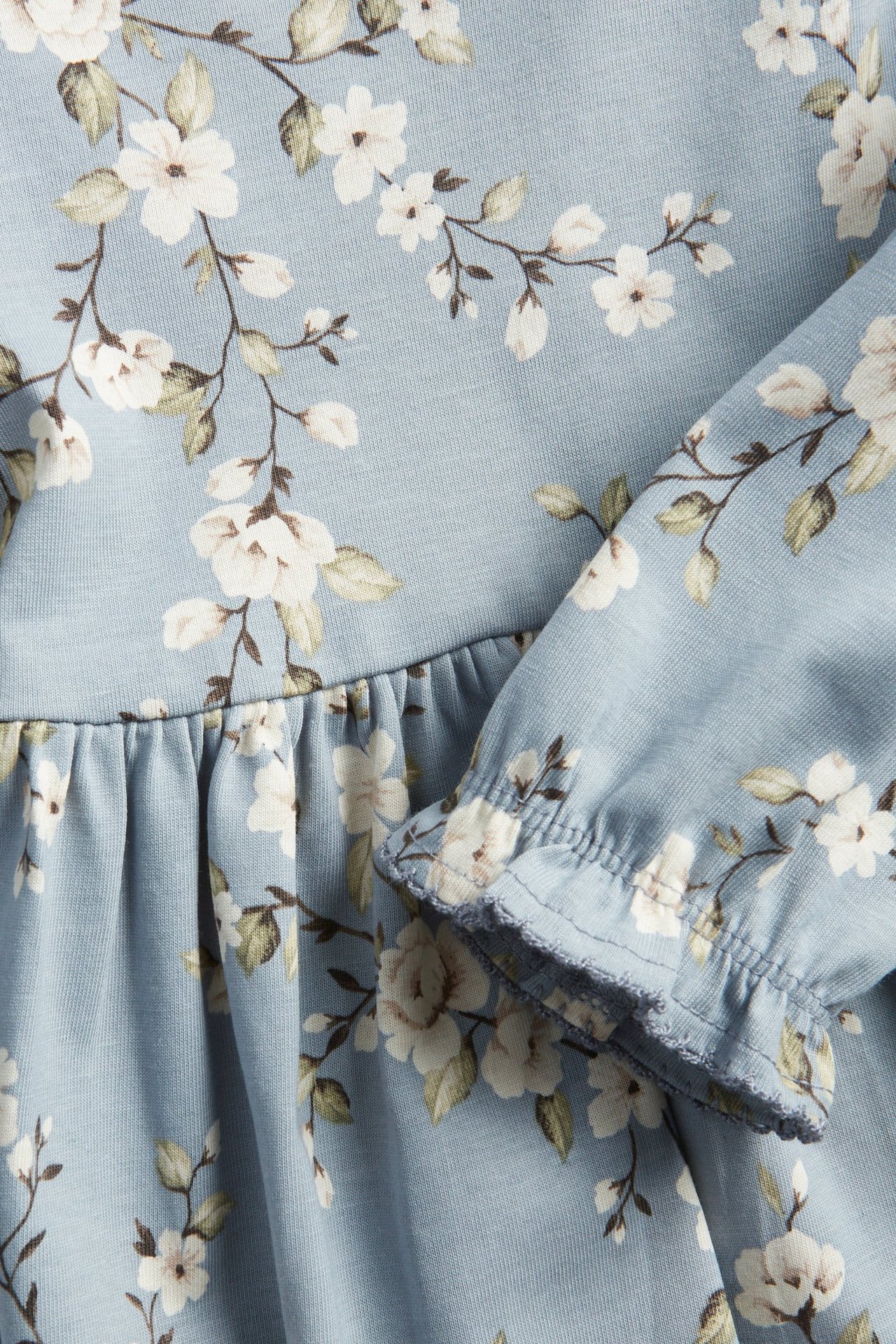 Zbliżenie na jasnoniebieską sukienkę z bawełnianej dzianiny w kwiaty, z marszczonym detalem i falbankowym mankietem.